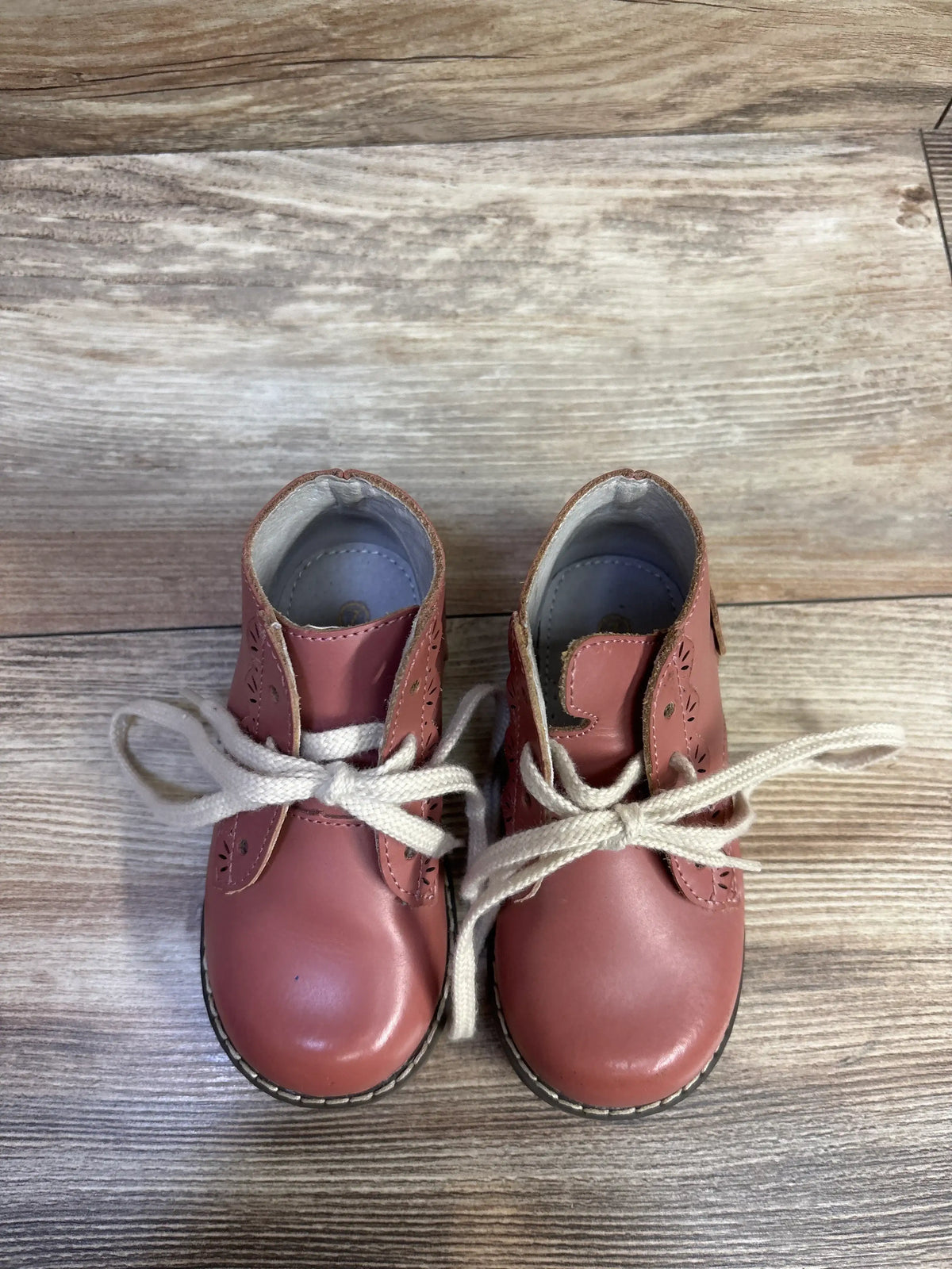 Top view of L’AMOUR Junie pink lace-up toddler boots showing brogue cutout detail and white laces on wood floor background