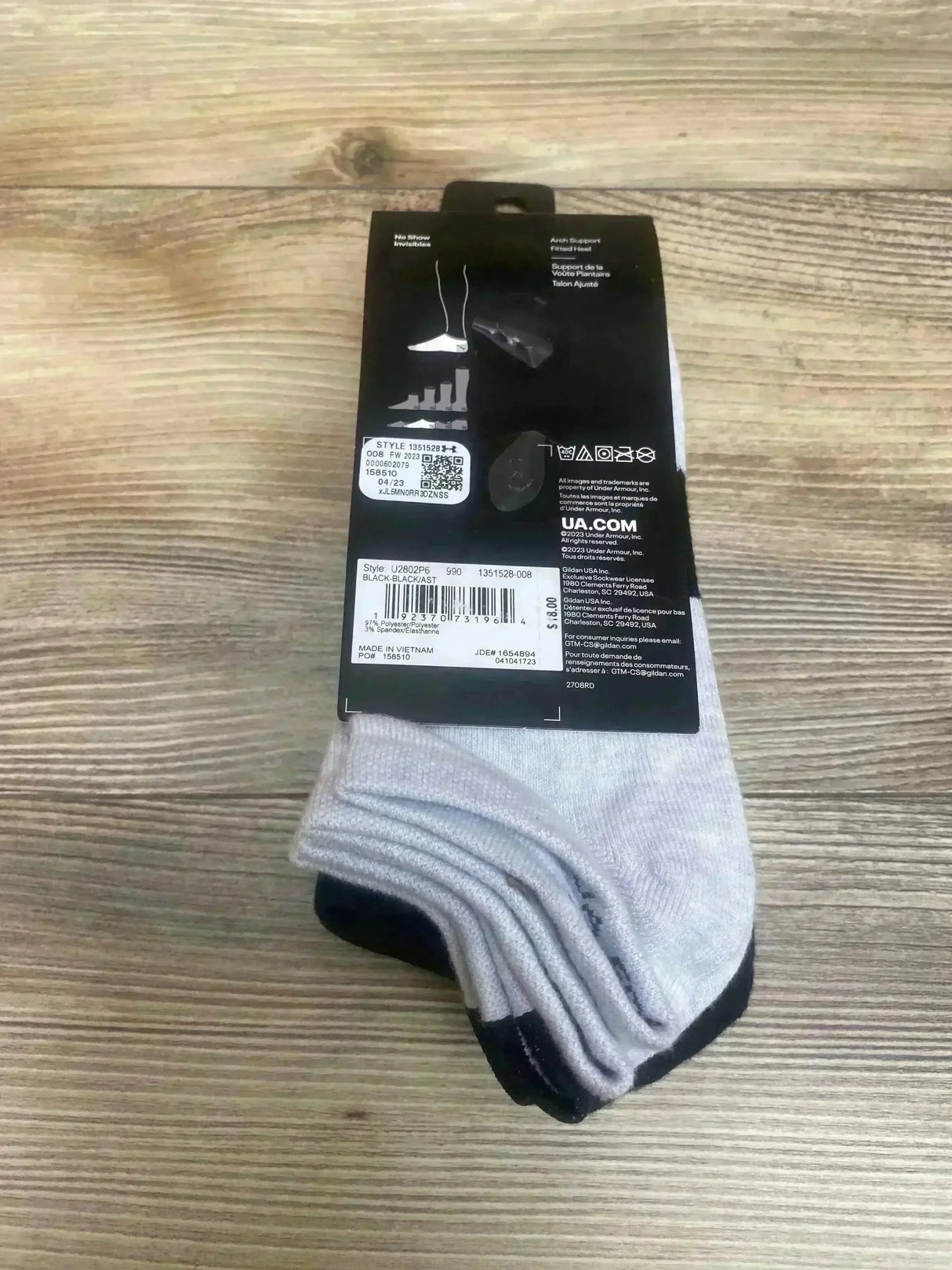 Top-down view of Under Armour six-pack kids no-show socks, light gray with black heels, folded under black label on wood floor
