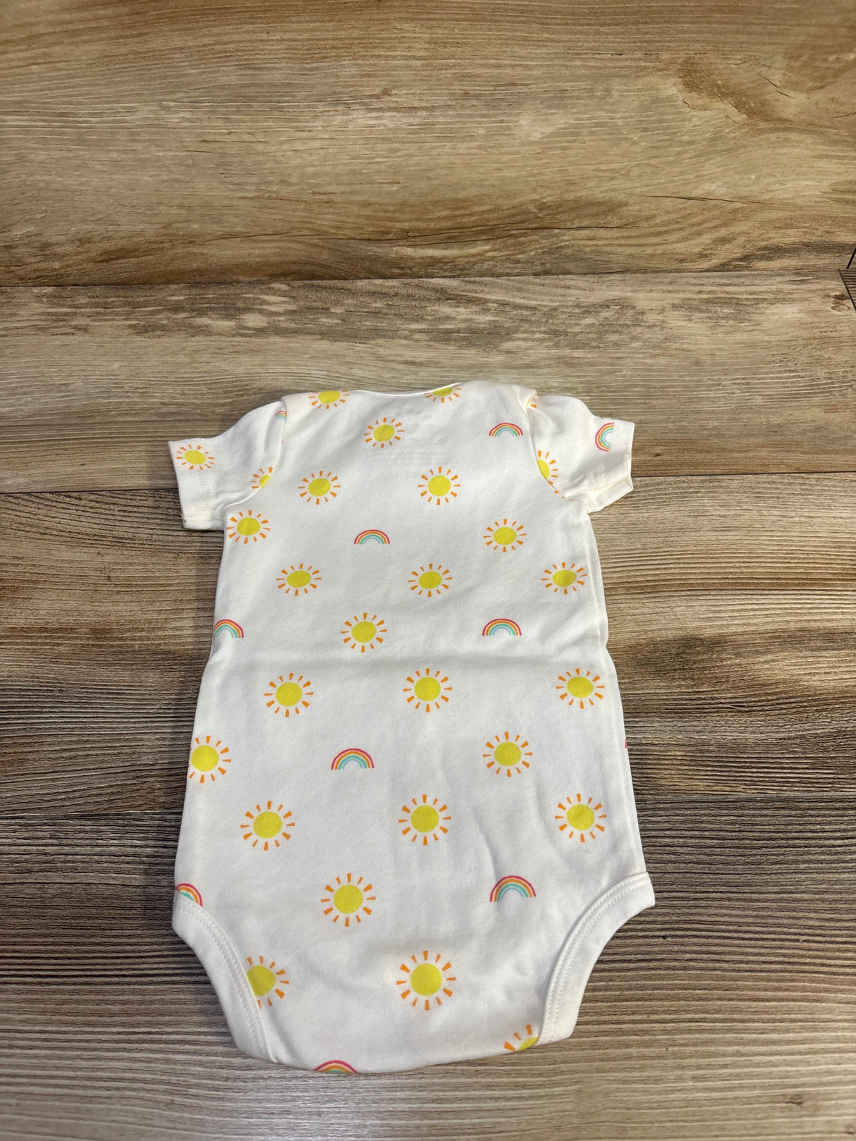 Flat lay of white sun-print baby bodysuit (3-6 months) showing yellow sun and rainbow prints on cotton wood background