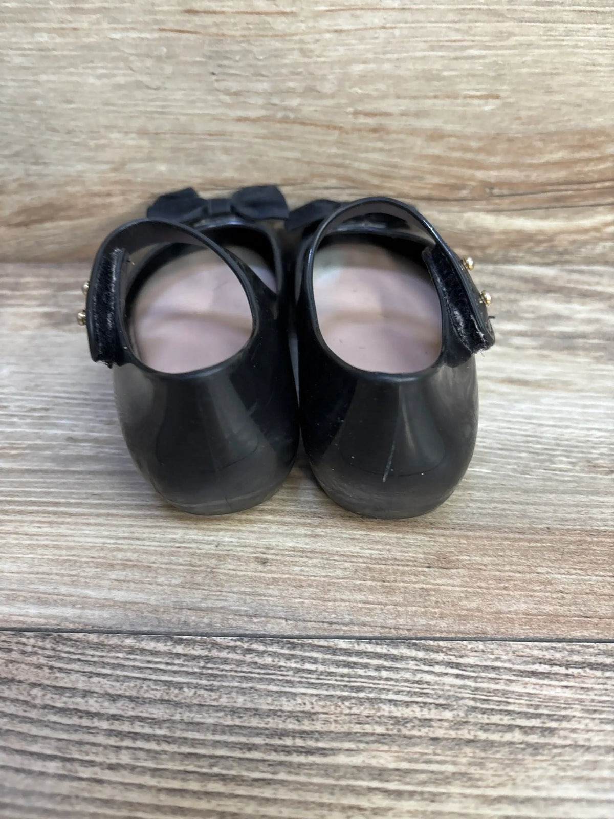 Rear view of Mini Melissa Ultragirl Bow jelly flats size 8 with back straps, gold studs, and bows on wood background