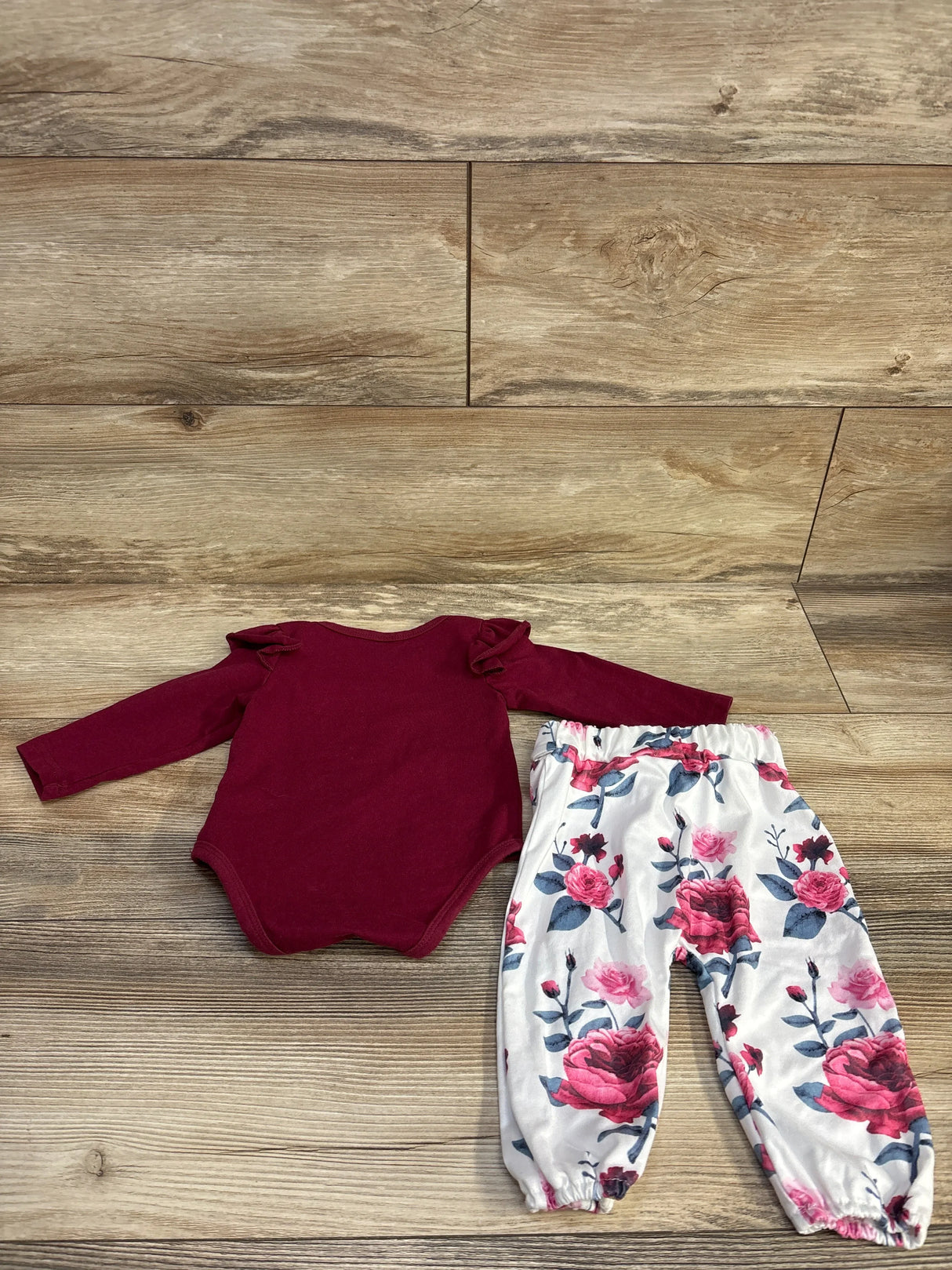 Burgundy baby bodysuit with ruffled shoulders and white floral pants on wood floor