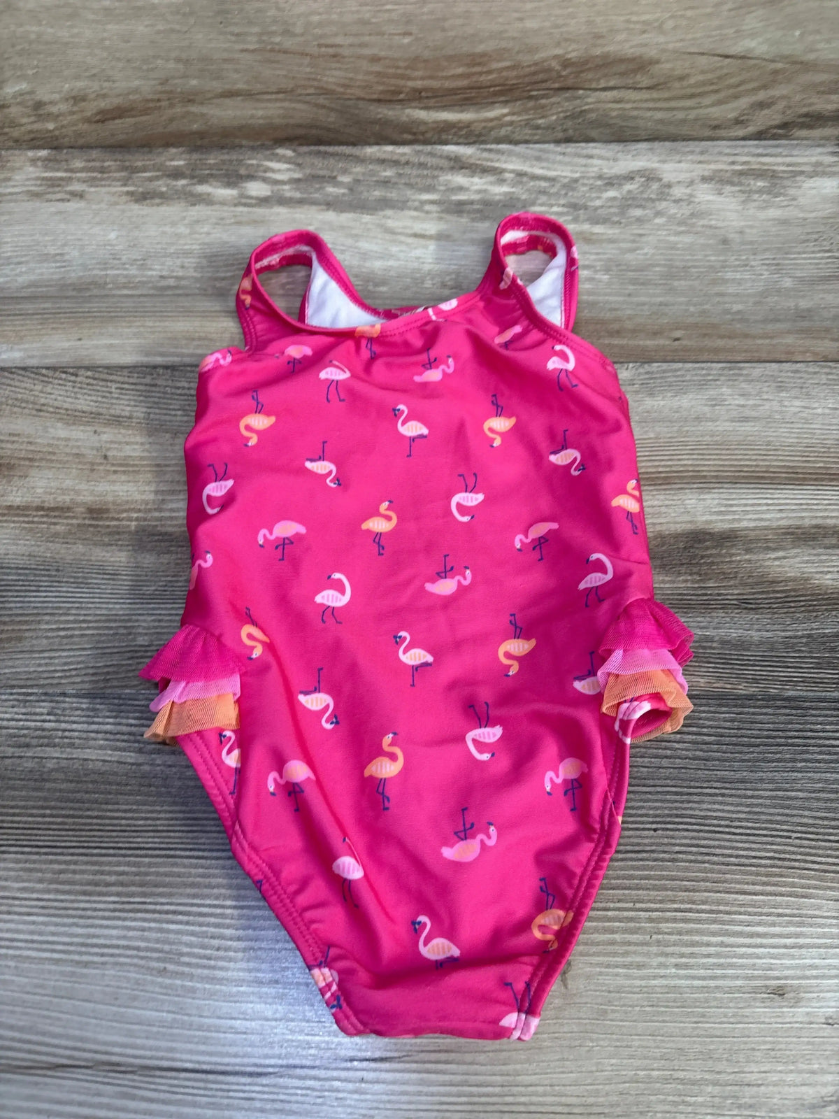 Flat lay of hot pink toddler one-piece swimsuit with small flamingo print and layered pink and orange tulle ruffle trim on light wood background
