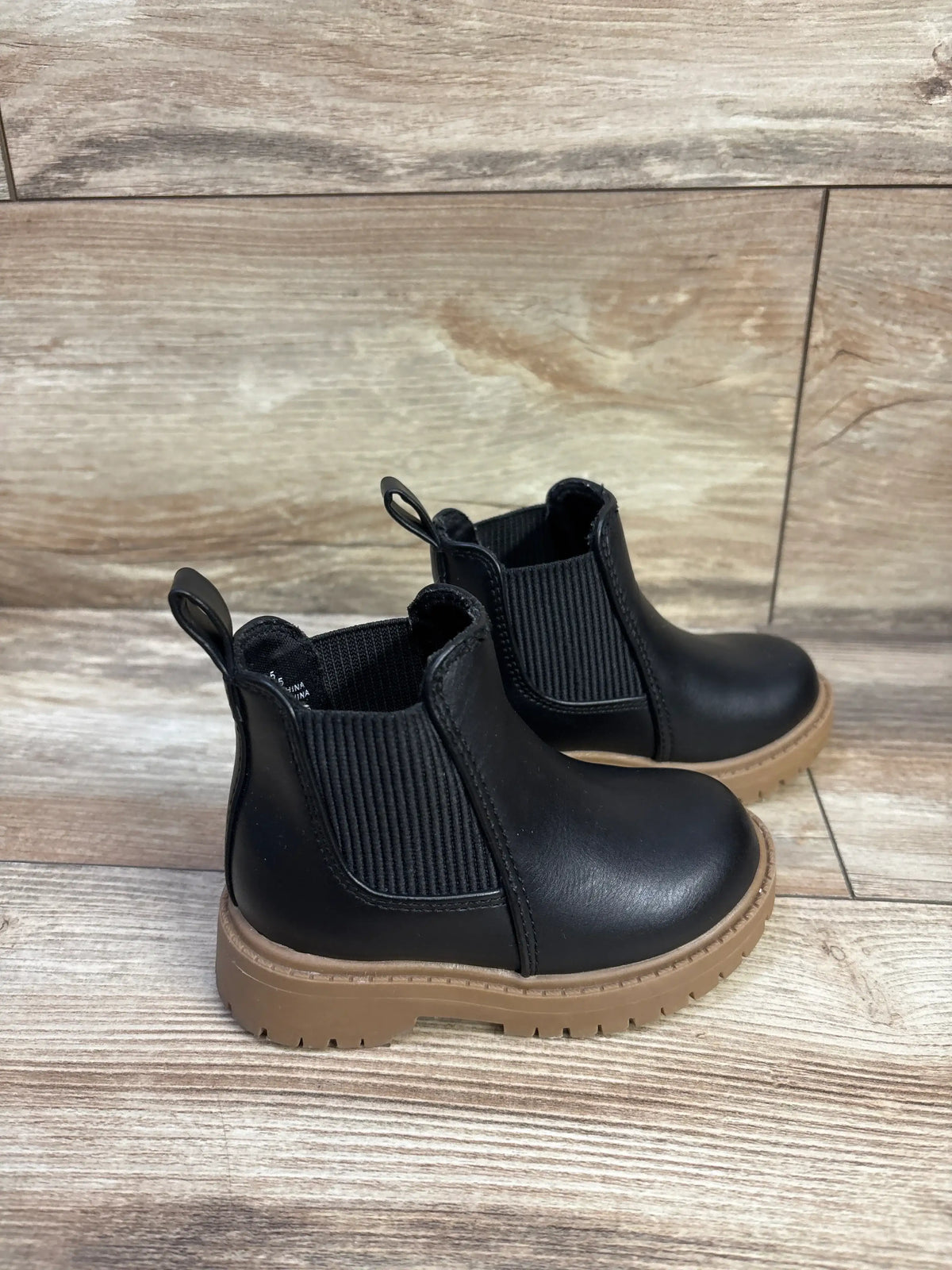 Side view of black faux leather toddler Chelsea boots size 5C with elastic side panels and tan lug sole on wood floor