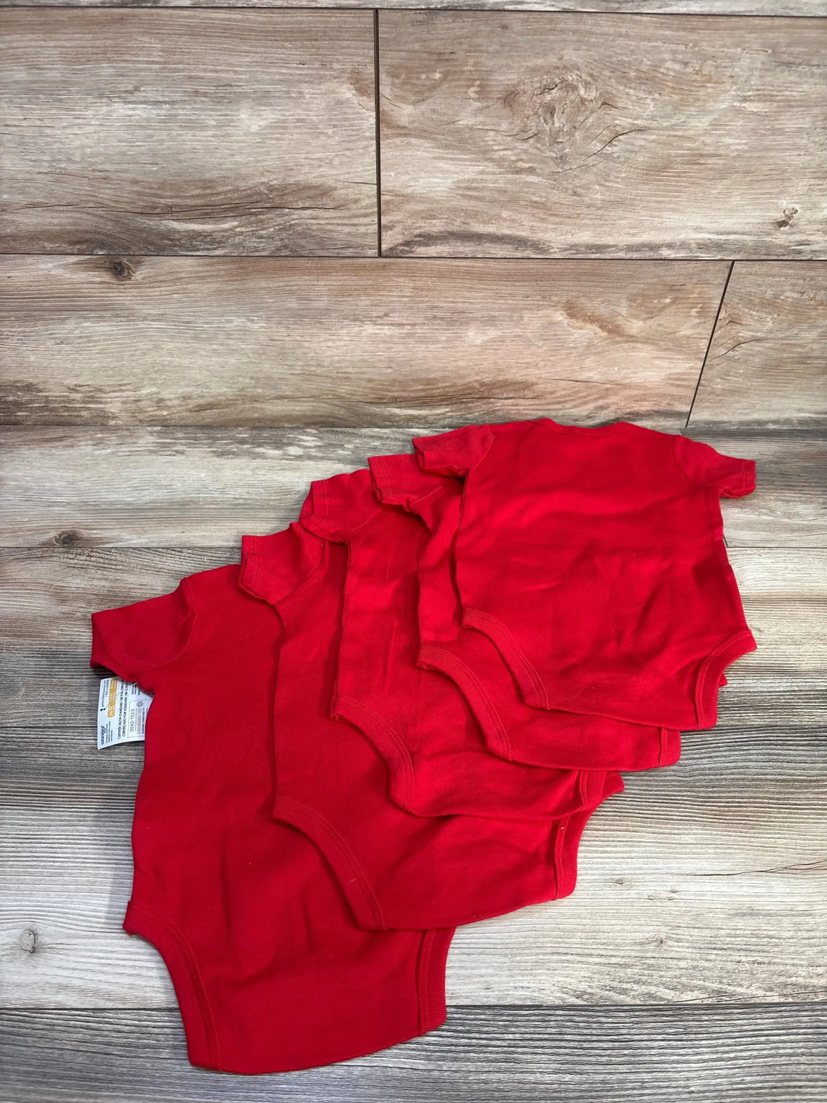 Flat lay of NEW Gerber 5pk bodysuit set in red showcasing fanned short-sleeve onesies with brand tags on wood plank floor