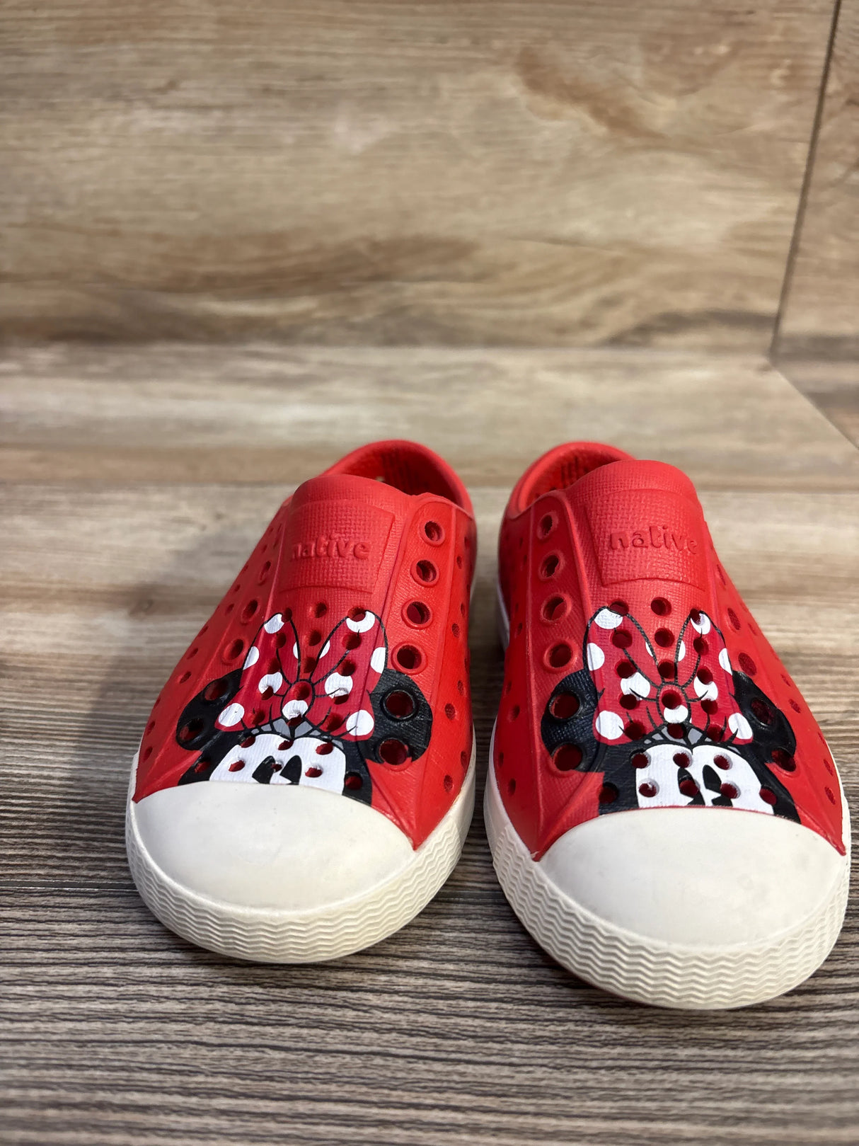 Front view of red Jefferson slip-on toddler shoes with painted Minnie Mouse faces and white toe caps on wood