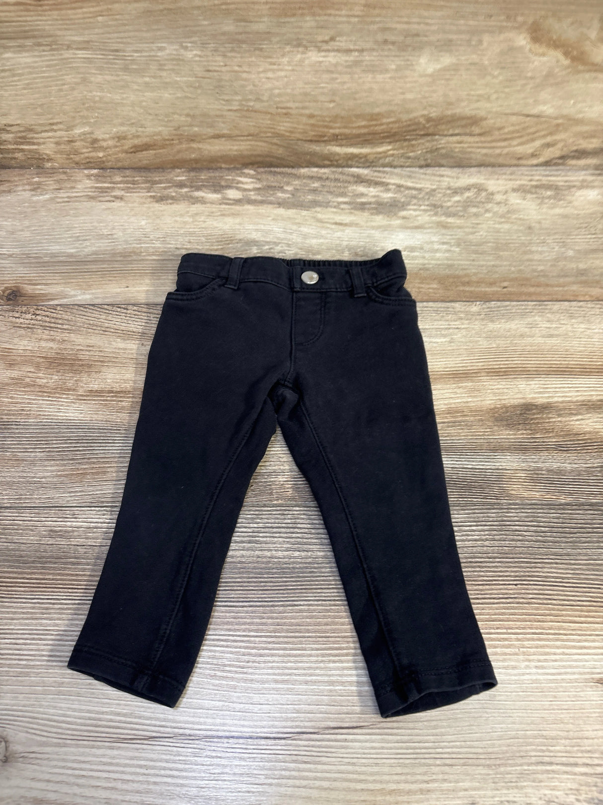 Front view of Children’s Place black toddler pants showcasing elastic waistband, faux button and slim tapered legs on wood background