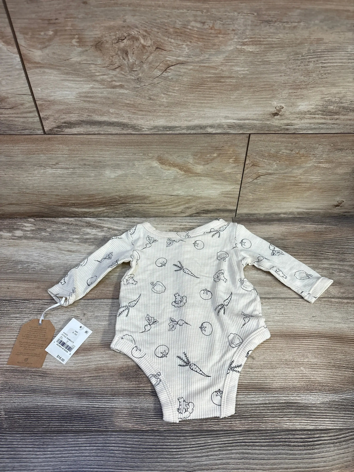 Flat lay of Grayson Collective ribbed ivory long-sleeve snap bodysuit (0–3M) showing vegetable line-drawing print and attached tags on wood floor