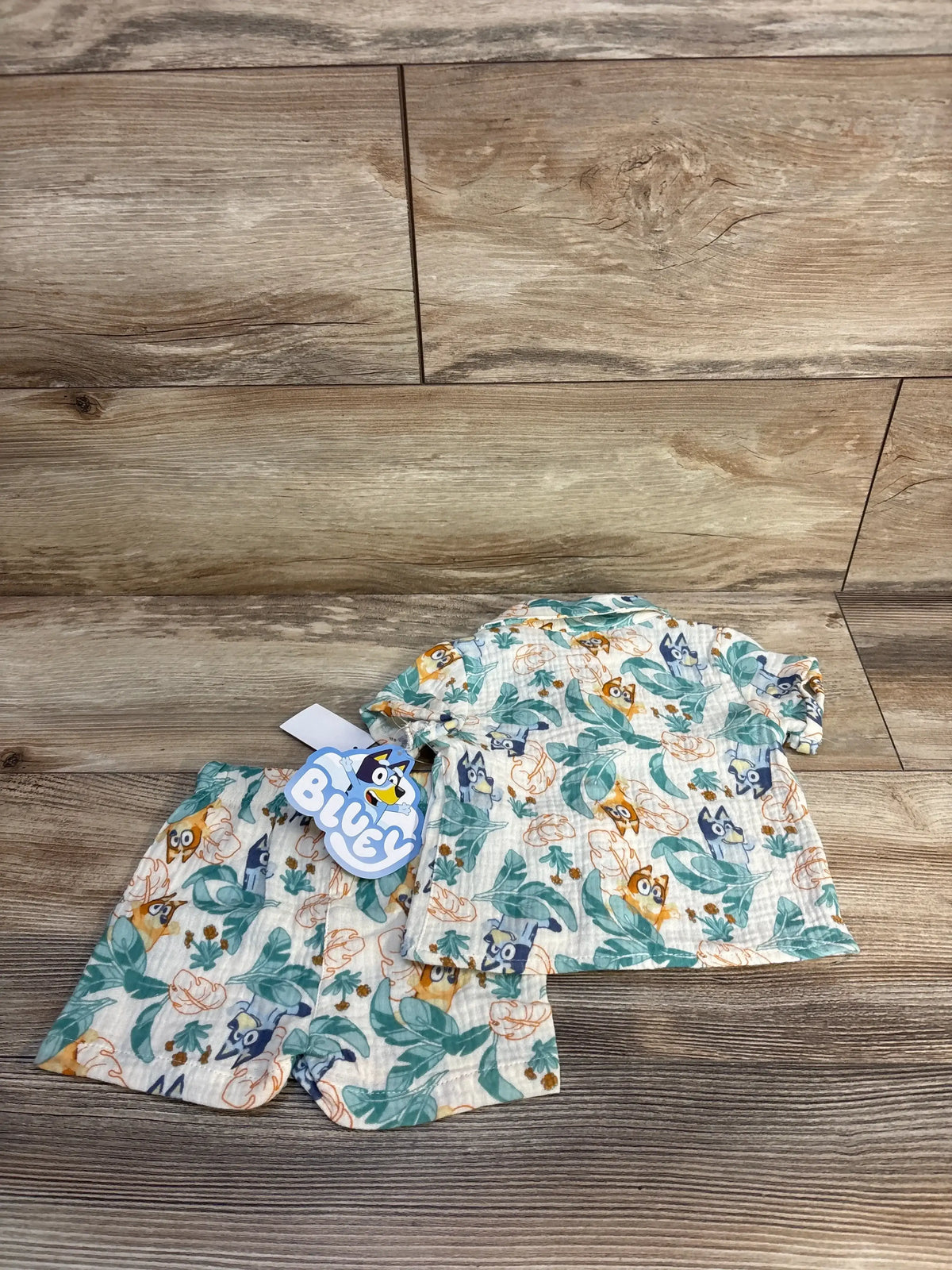 Top-down view of cream Bluey 2-piece button-up gauze shirt and shorts set showcasing leaf and character print on wood floor