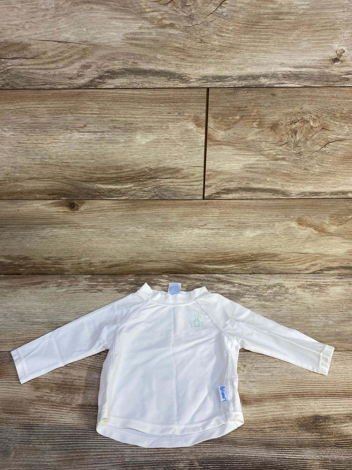 Front flat lay of white iPlay baby rashguard showing crew neck, mint chest logo and brand tag on wood background