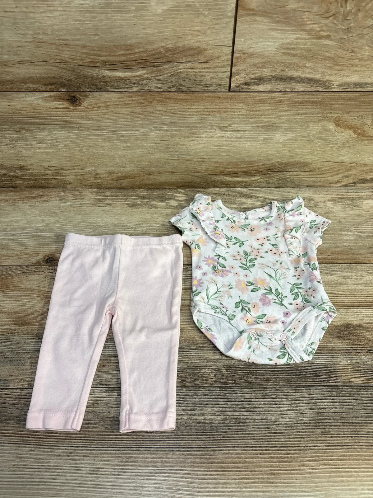 Flat lay of 2-piece baby set: floral ruffle-sleeve bodysuit with snap crotch and light pink pants, size 3M on wood background