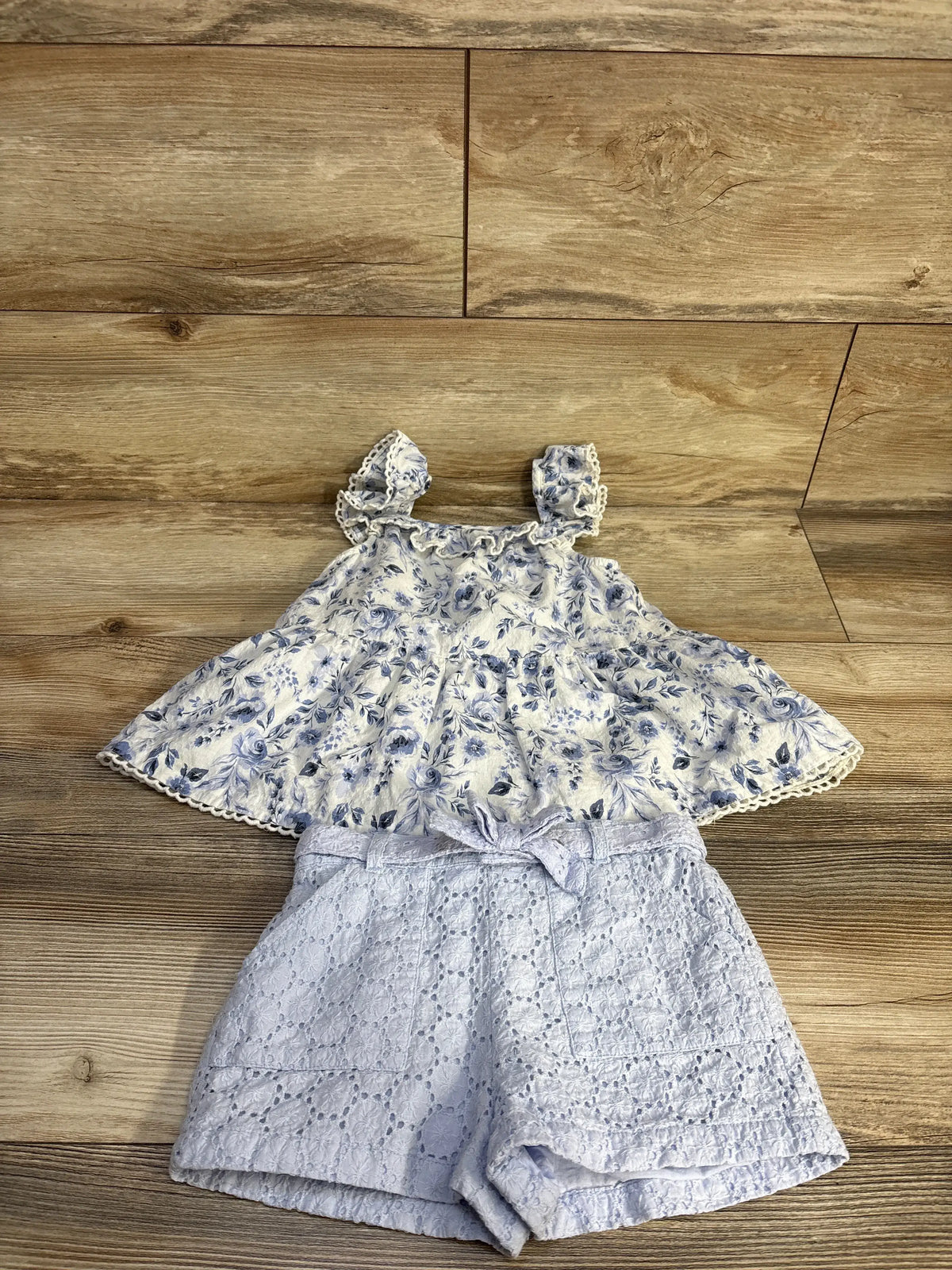 Flat lay of Joie blue floral blouse and lace short set with sleeveless 100% cotton top, belt, and lace shorts on wood floor