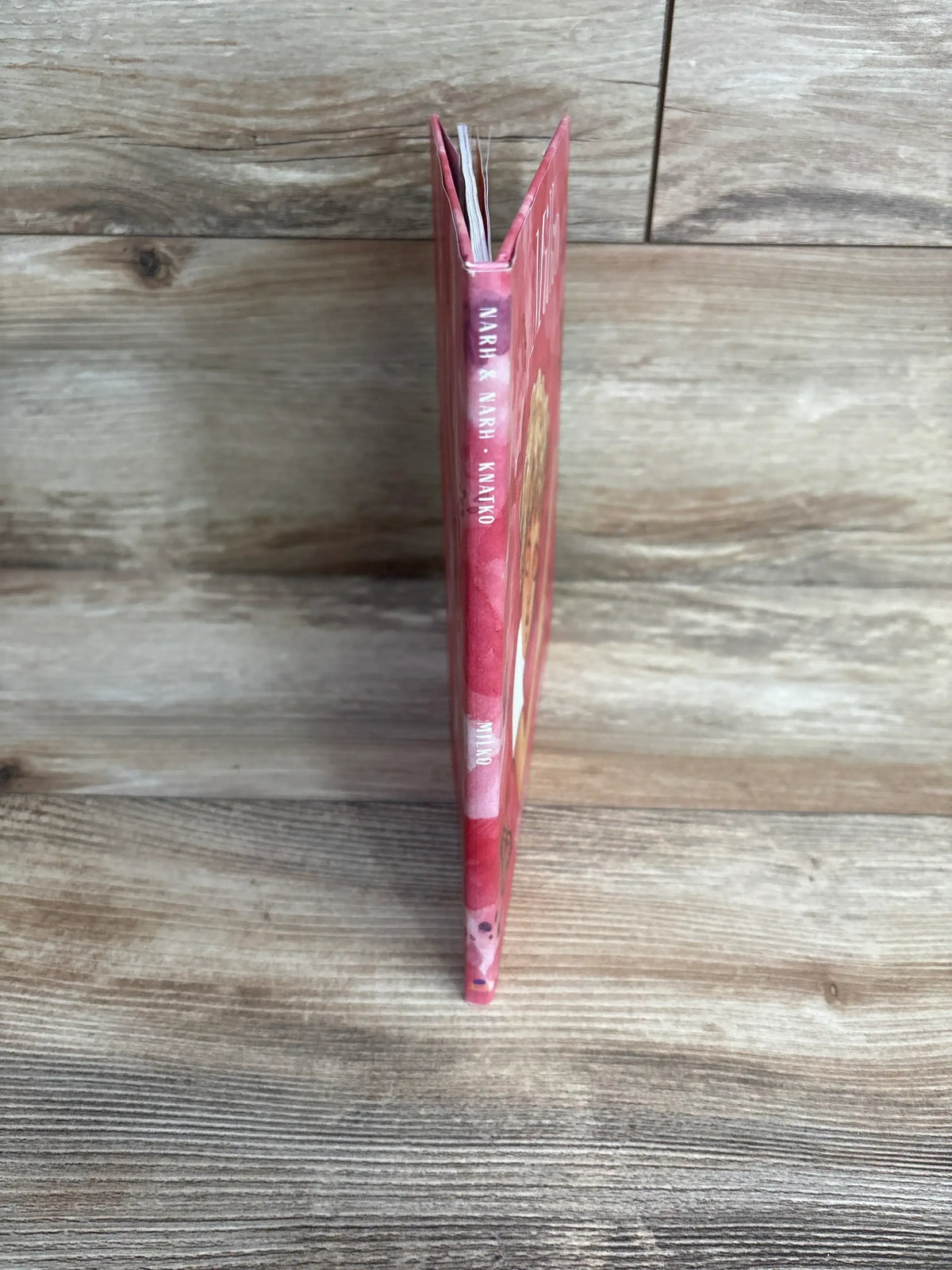 Spine view of Milko Hardcover Book with pink cover standing upright on wood plank floor
