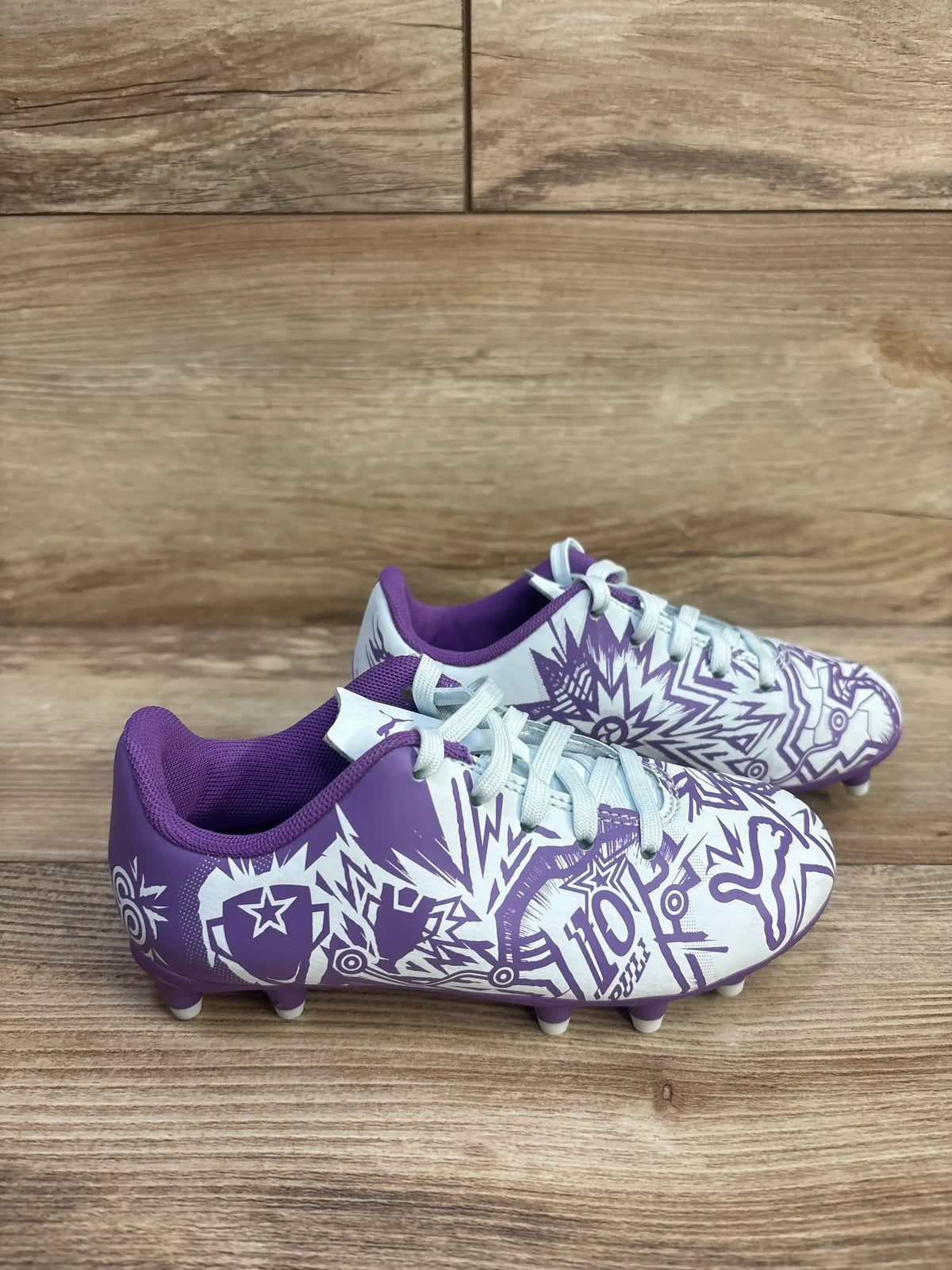 Side view of purple and white Puma x Christian Pulisic Tacto II soccer cleats with graphic uppers and studs on wood