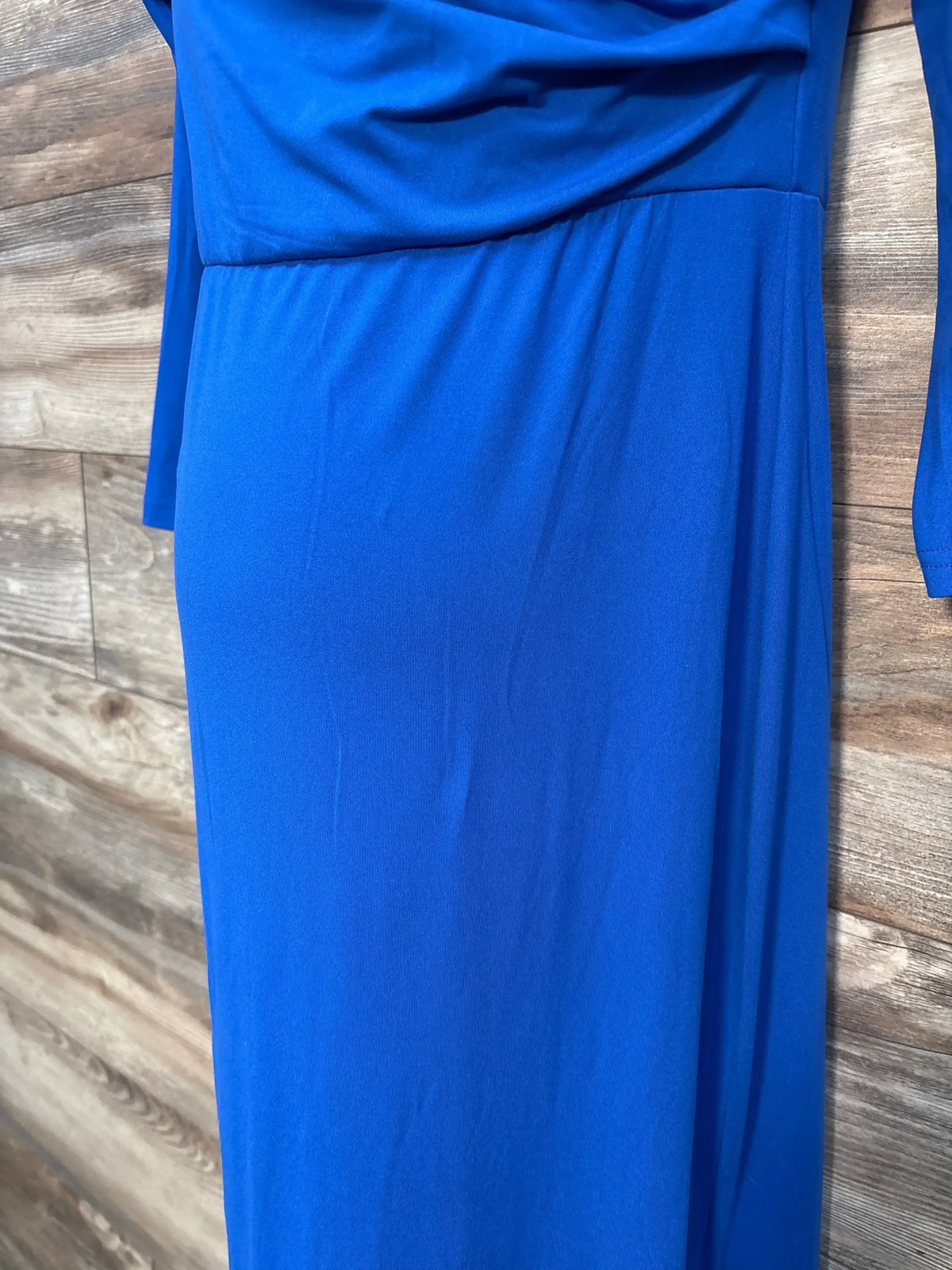 Close-up of maternity off-shoulder royal blue dress showing draped neckline, waist seam and long sleeve against wood-paneled background