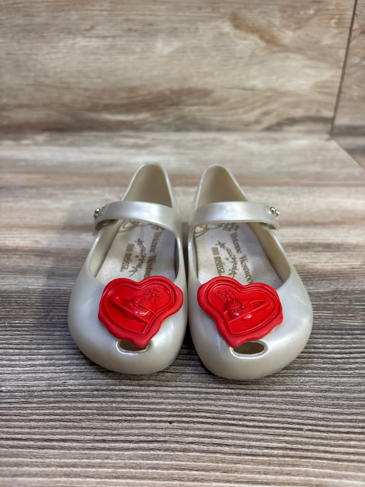 Front view of Vivienne Westwood x Mini Melissa Ultra shoes, pearl ivory Mary Janes with red heart orb charms on wood backdrop