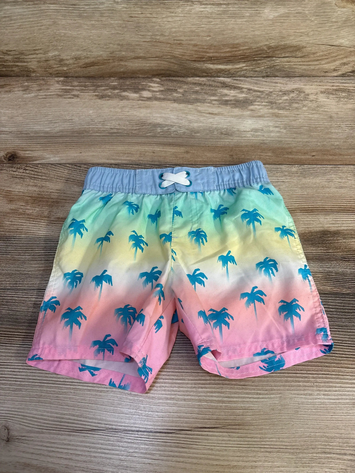 Front view of Cat & Jack palm tree swim trunks, size 4T, showcasing elastic waistband and drawstring on wood-grain background