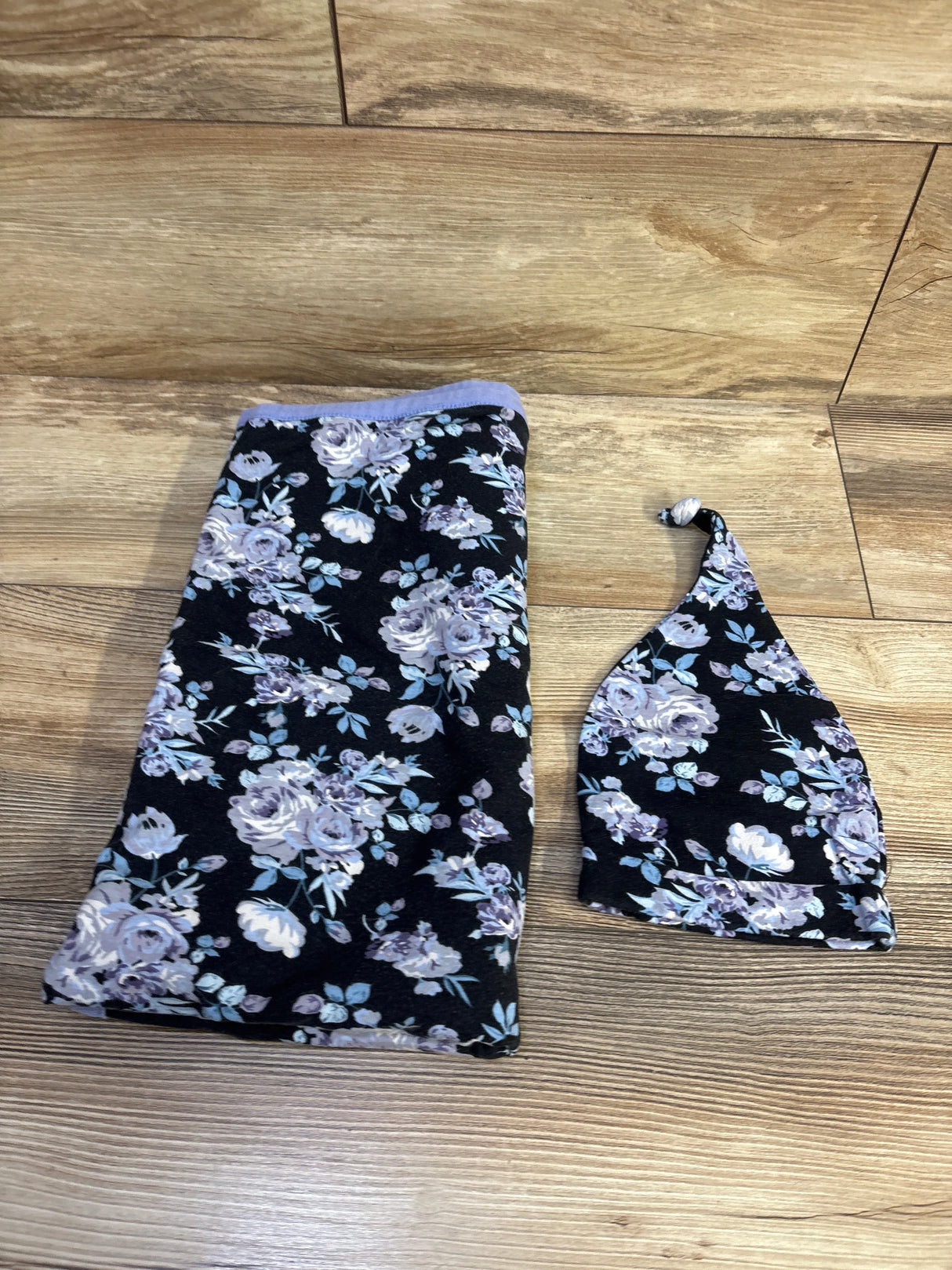 Flat lay of 2-piece floral swaddle blanket and knotted hat showing lavender and white roses on black fabric on wood floor