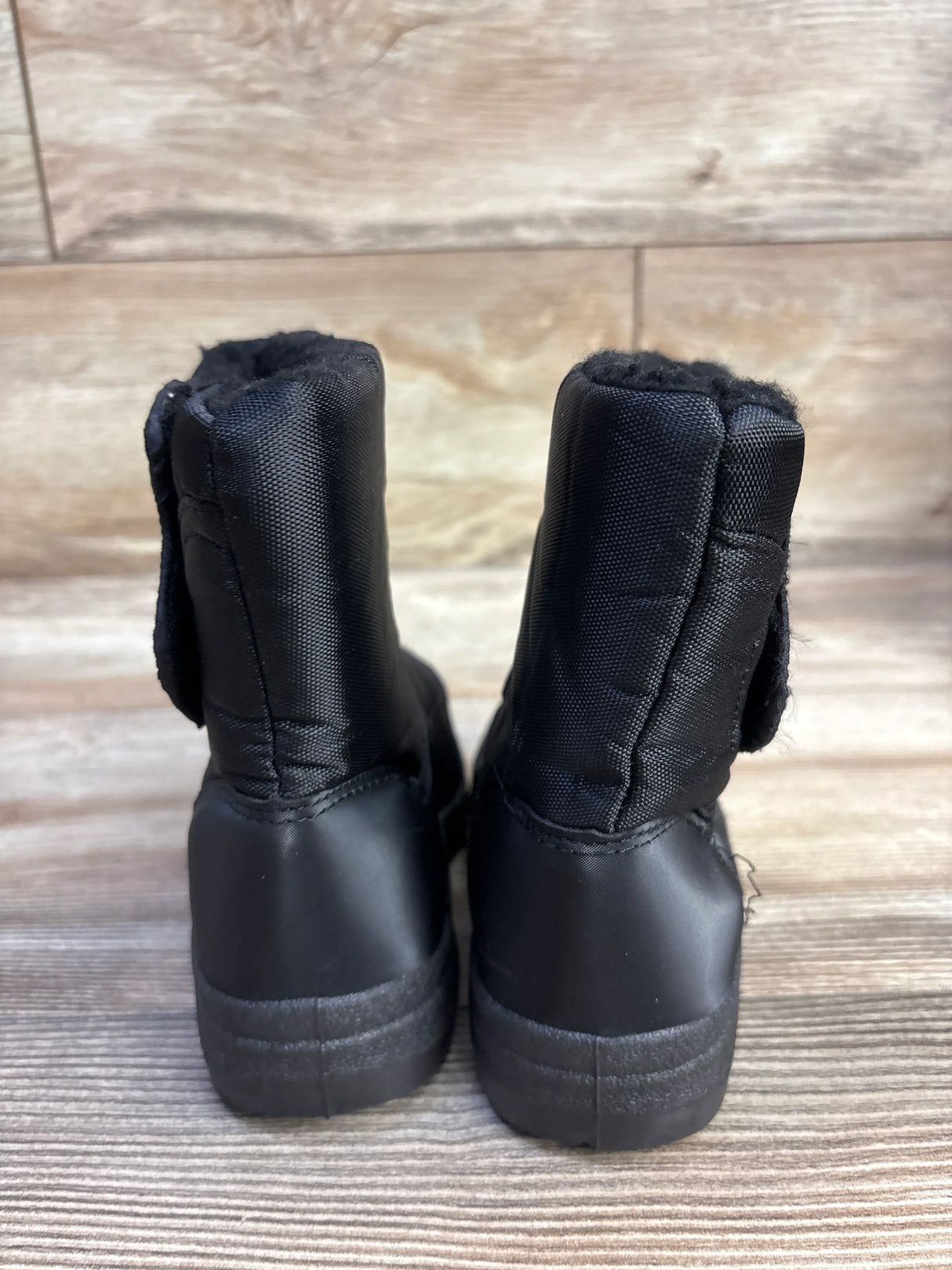 Rear view of toddler black snow boots showing plush fur lining, side hook-and-loop strap, and textured rubber soles on wood floor