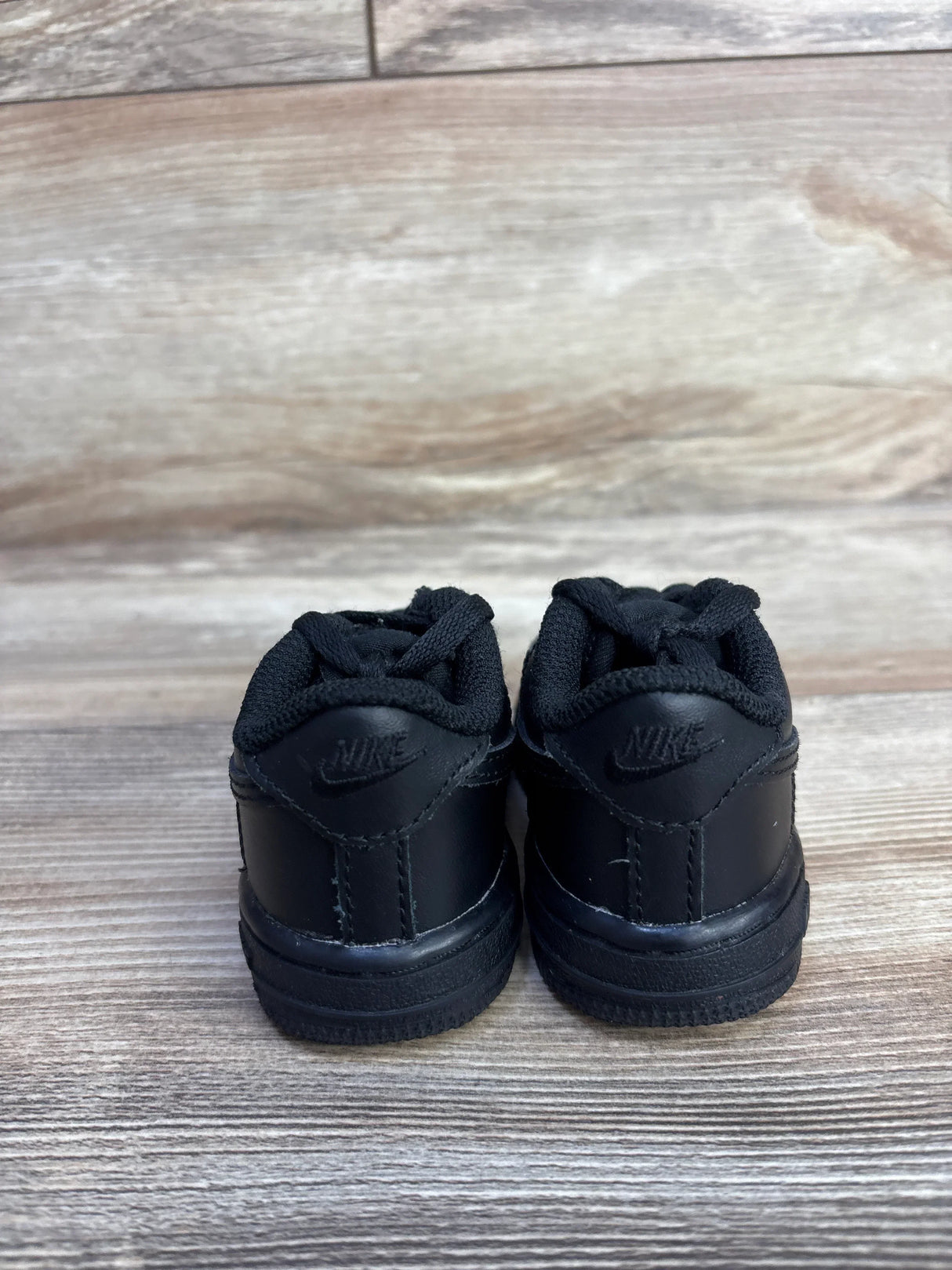 Back view of Nike Air Force 1 06 TD Triple Black Sneakers showcasing all-black leather heel with embossed Nike logo on wood plank floor