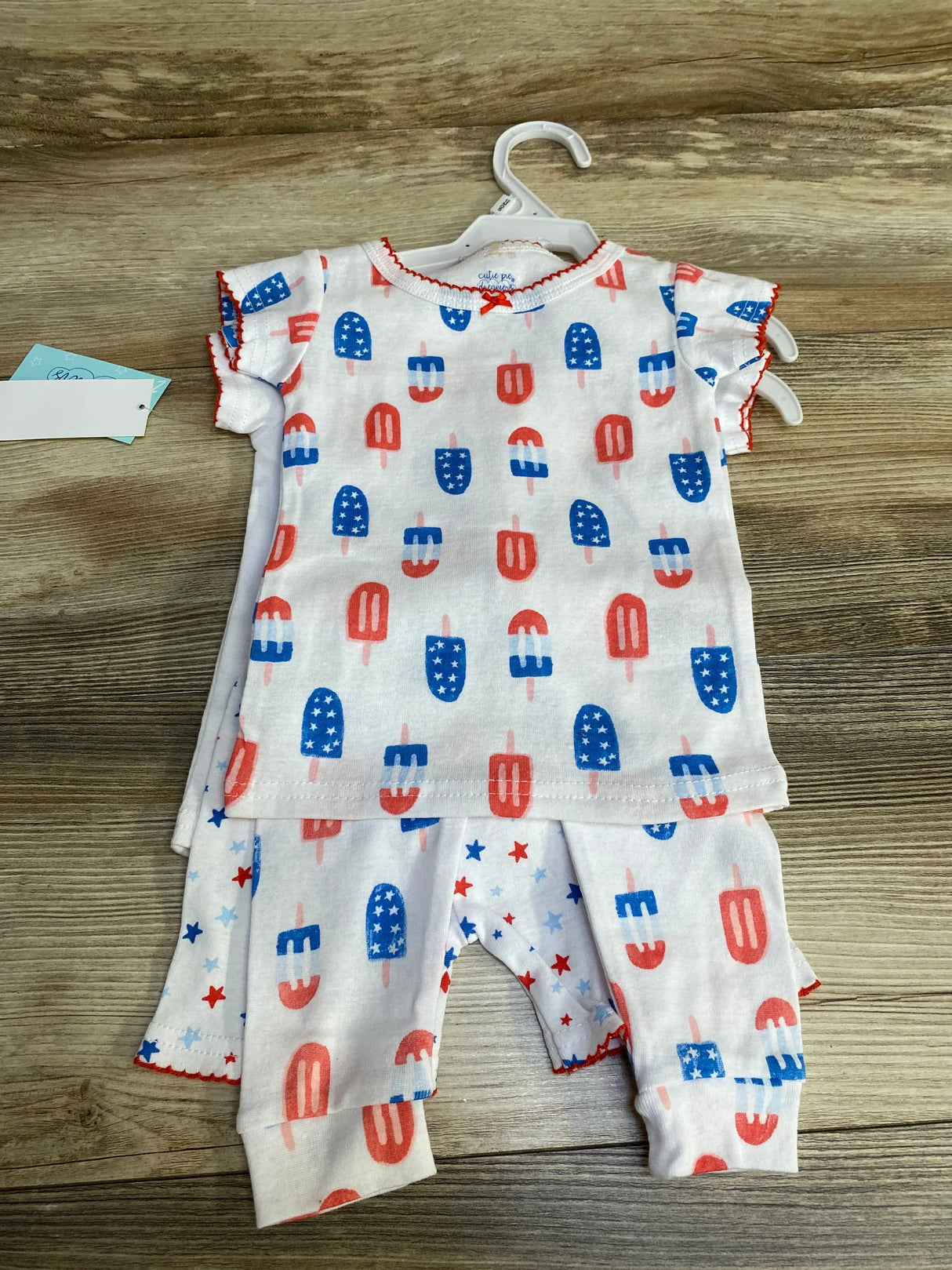 Flat lay of 4-piece white baby pajama set (size 12M) showing red and blue popsicle and star print on wood background
