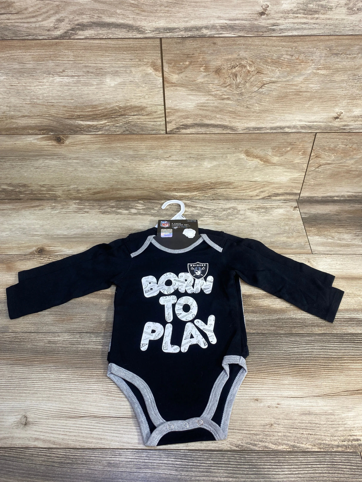 Black Raiders baby onesie with 'Born to Play' text, laid on wood floor