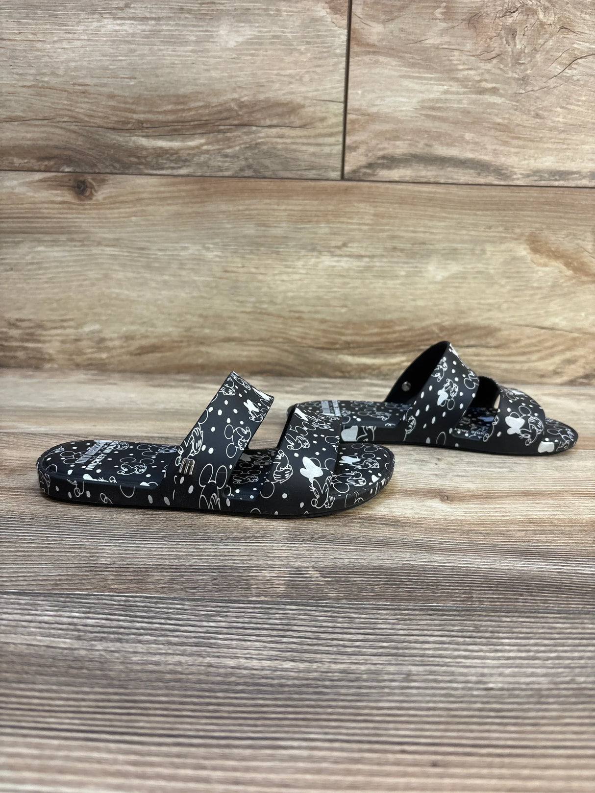 Three-quarter view of Mini Melissa x Disney Minnie Mouse Beach Slides in black showcasing white Minnie Mouse character print and dual straps on wood plank floor