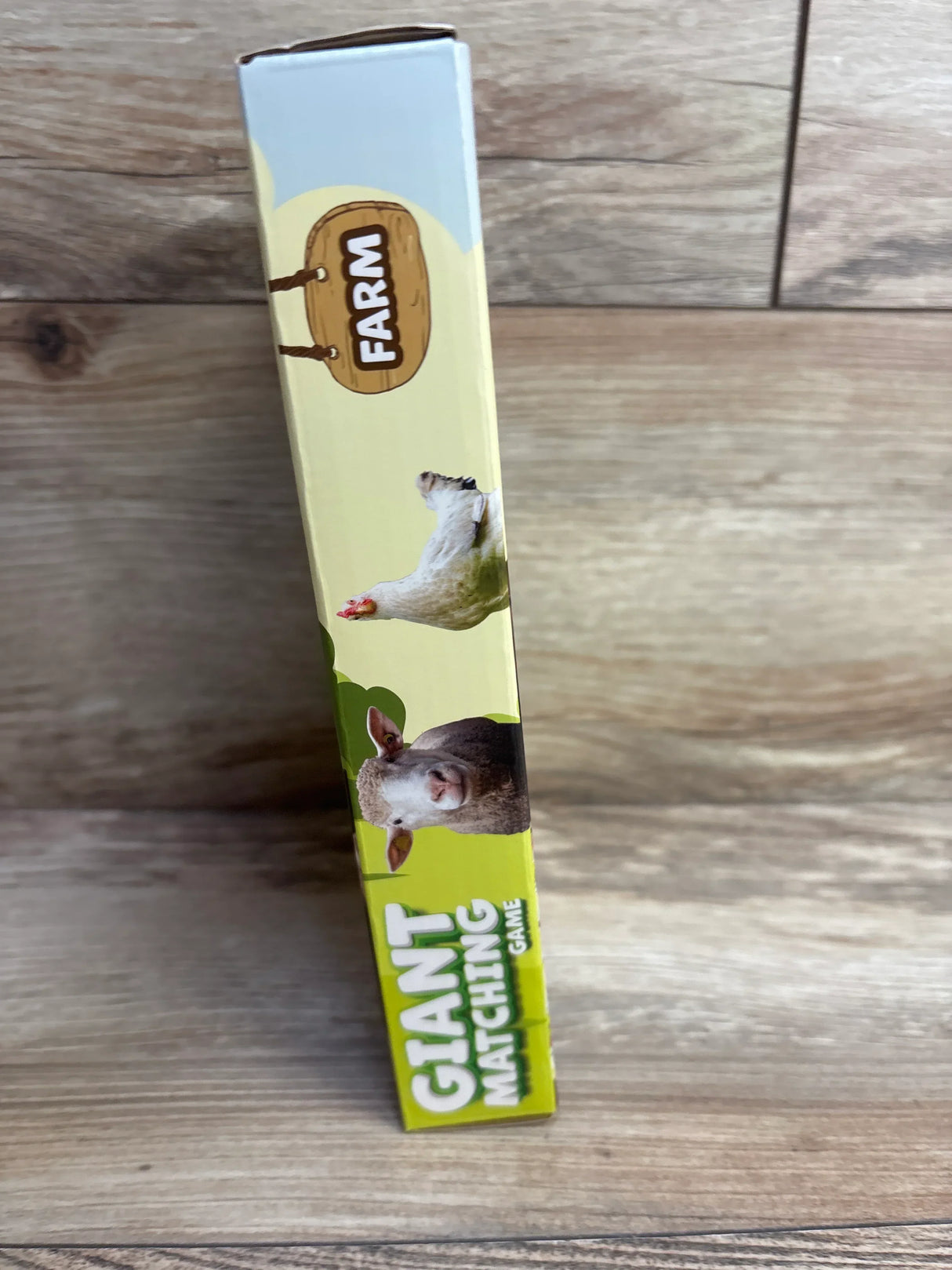 Angled side view of PlayMonster Giant Matching Card Game box (Farm) showing FARM sign and sheep and chicken illustrations on wooden background