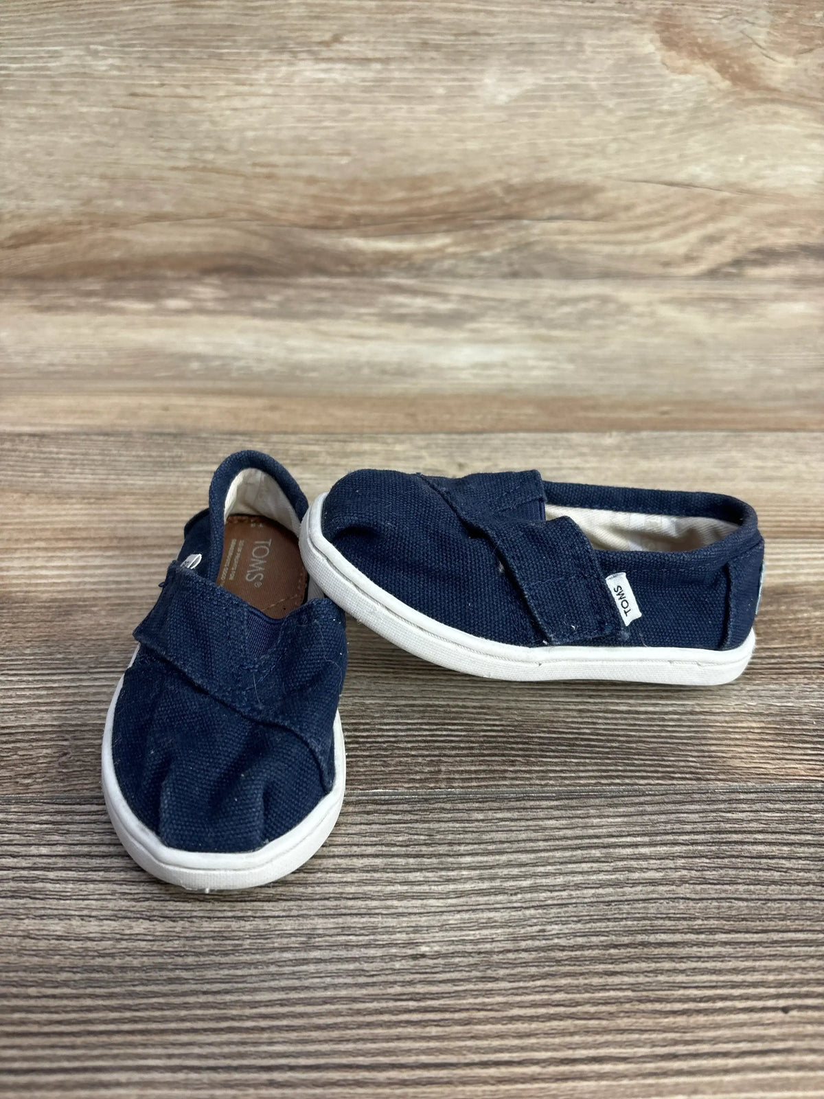 Angled view of Toms Tiny Alpargata navy slip-on (5C) showing canvas upper, Velcro strap and white sole on wood background