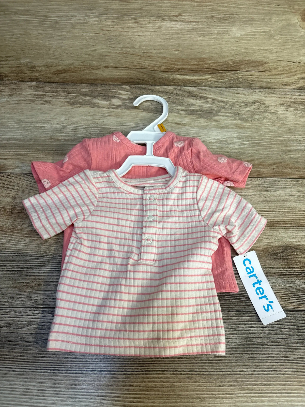 Two infant girls’ Carter’s shirts on hangers, one pink striped and one solid pink with daisies, on wood background.