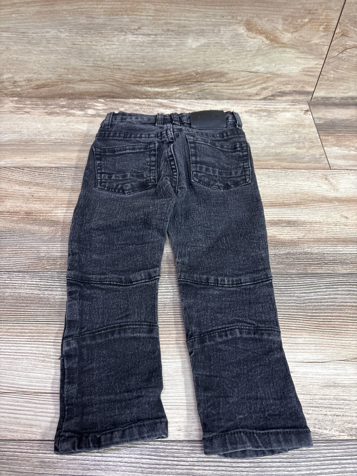 Back view of Steve’s Jeans black moto jeans showcasing rear pockets and paneled legs on a wood-look floor