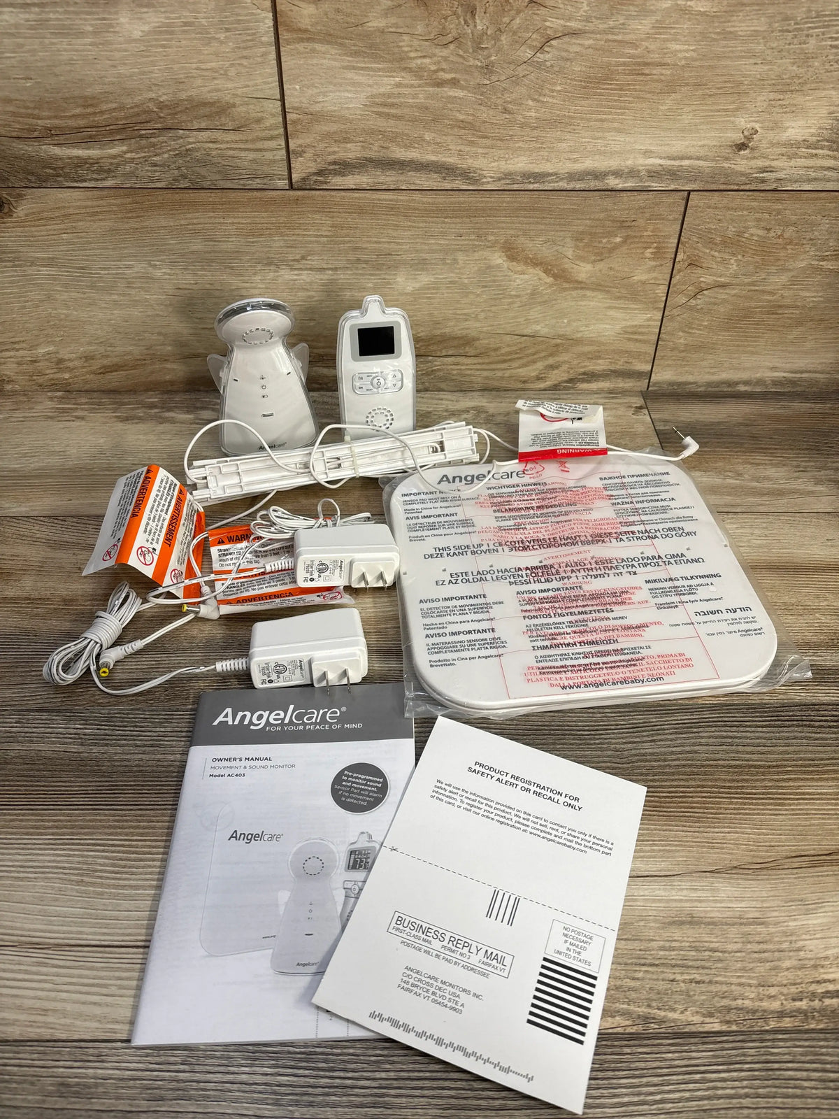 Angled overhead of Angelcare Movement and Sound Monitor showing parent and baby units, sensor pad, and manuals on wood floor