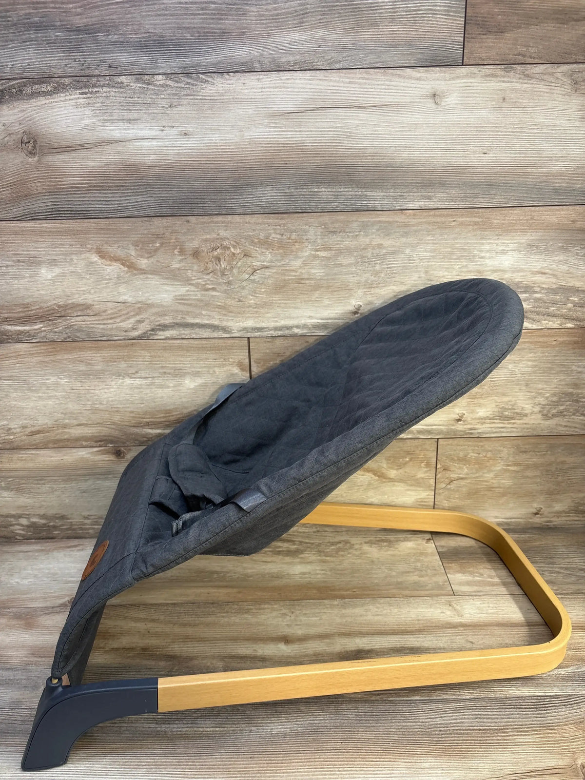 Side view of ANGELBLISS baby bouncer in grey with quilted seat, safety harness, and curved wood base on wood backdrop