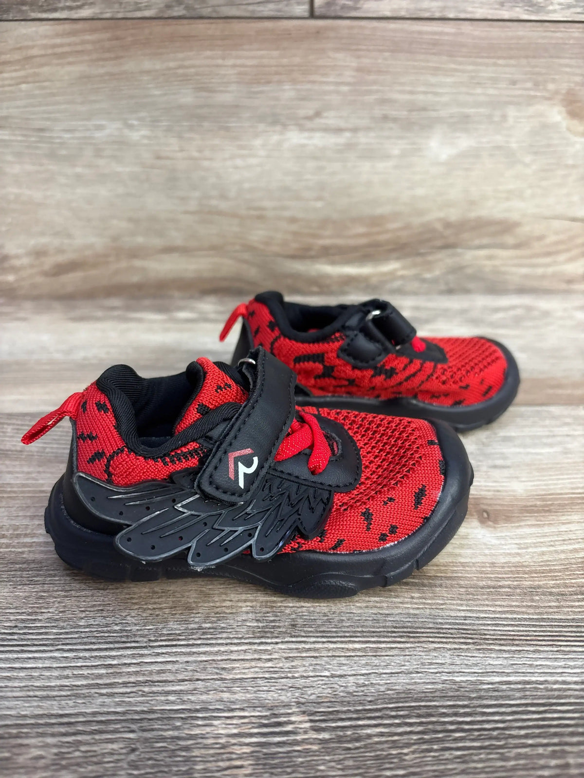 Side view of boys' red and black wing sneakers showing knit upper, wing-shaped Velcro strap, and black rubber sole on wood background