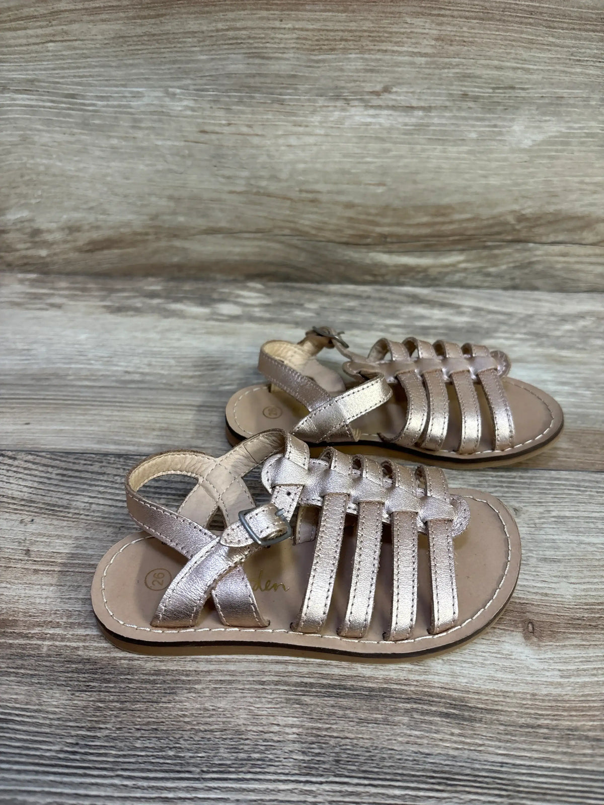 Front-side view of Mini Boden gold toddler gladiator sandals showing metallic leather straps and ankle buckle on wood background