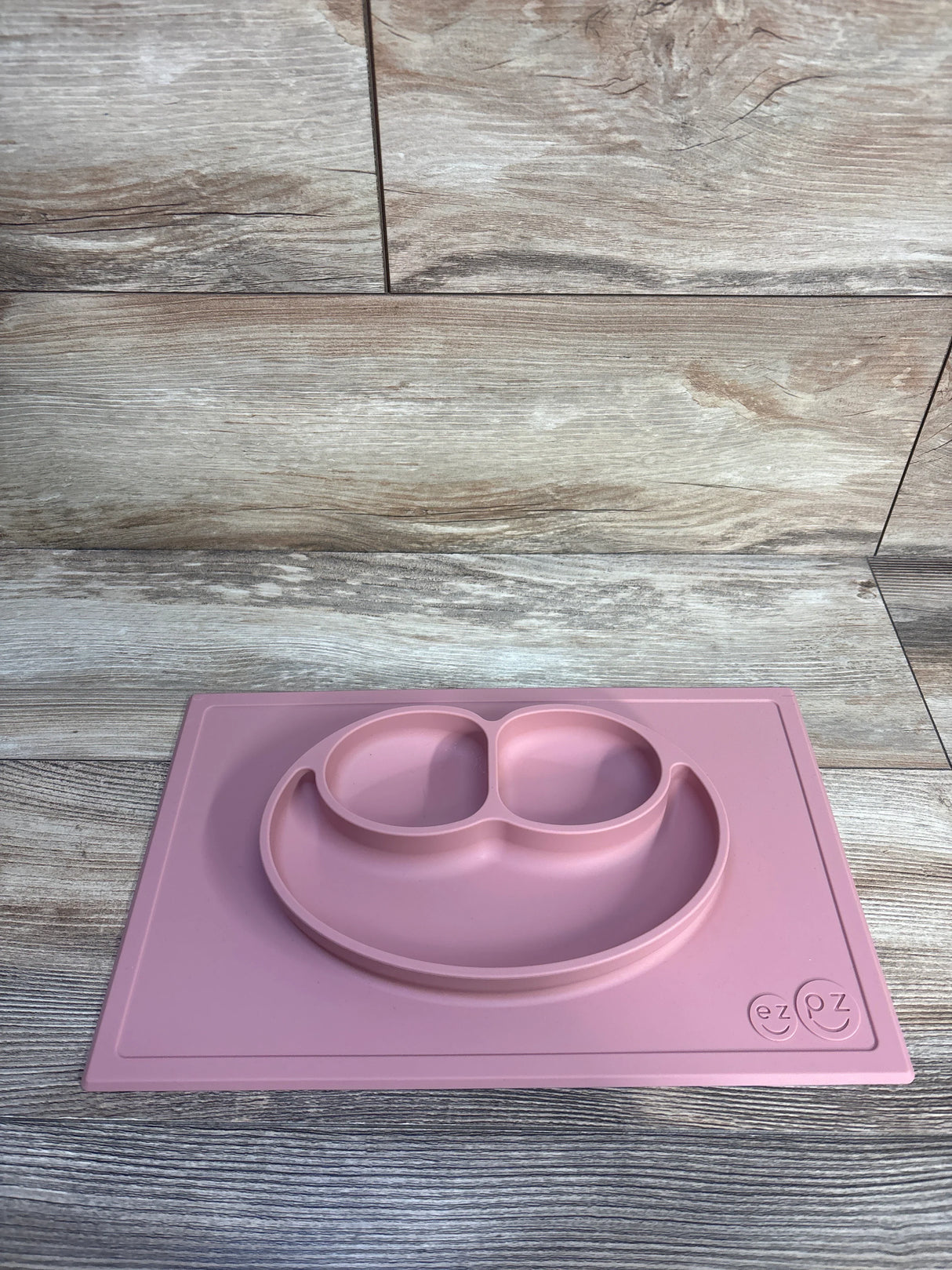 Top down view of ez pz Happy Mat suction plate and placemat in blush showing three compartment smile plate on wood background