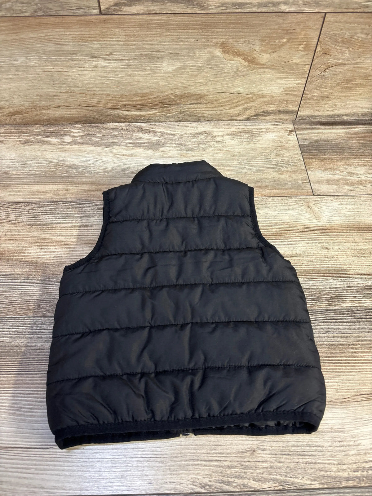 Back of black Puma puffer vest, size 12M, showing horizontal quilted panels and stand collar on light wood floor