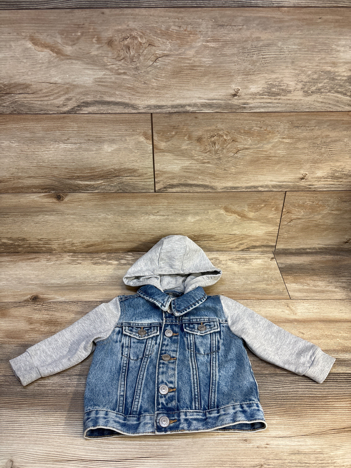 Baby denim jacket with gray hoodie sleeves, laid flat on wood floor
