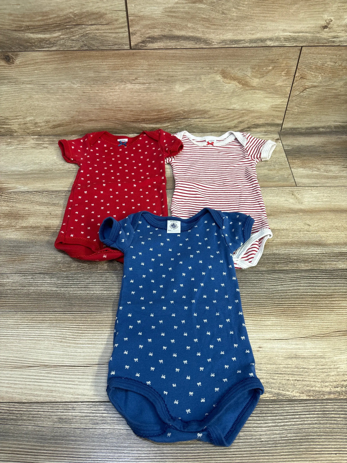 Top view of 3 Petit Bateau 6M short-sleeve bodysuits, red, red-and-white stripe, and navy with white bow print on wood floor