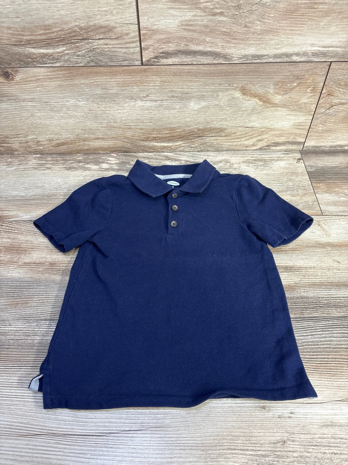 Front view of a navy Old Navy polo shirt in size 5T showcasing its collar, three-button front, and short sleeves on a wood-look floor