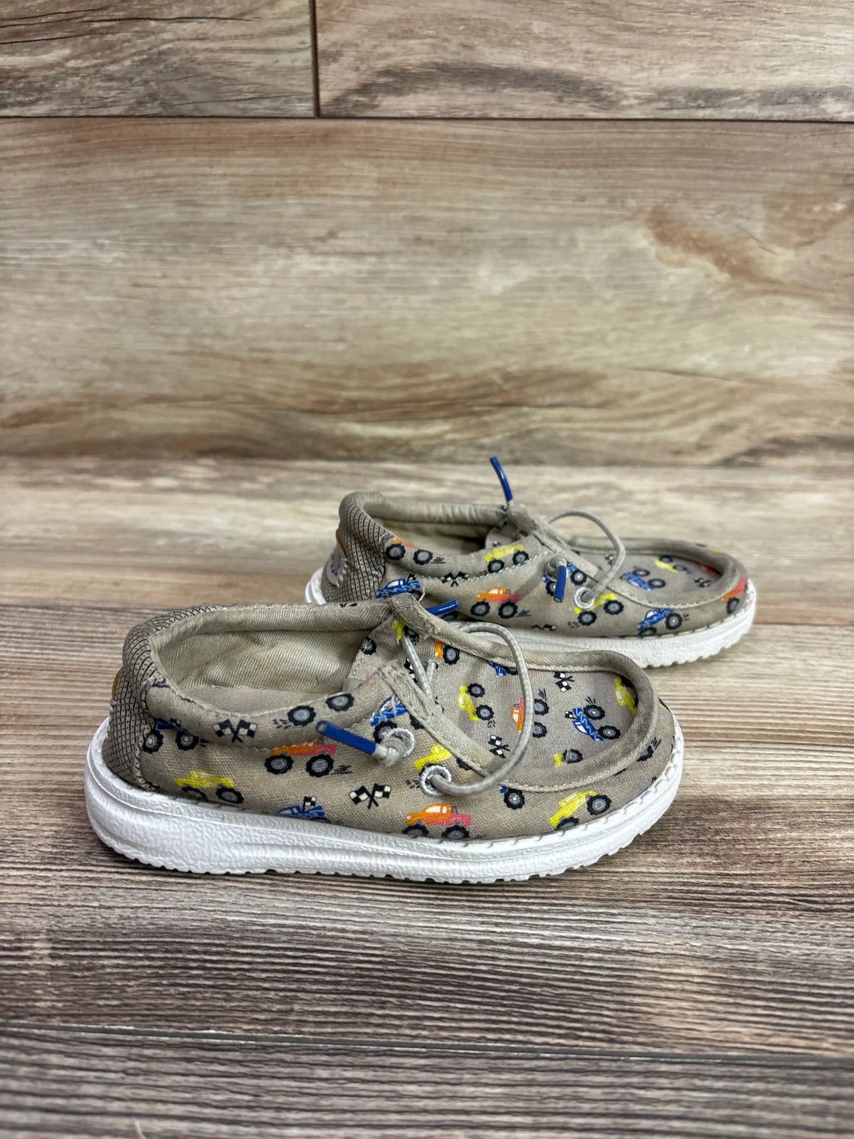 Side view of Hey Dude Wally T Trucks shoes showcasing tan fabric, colorful vehicle prints, laces, and white soles on a wood surface