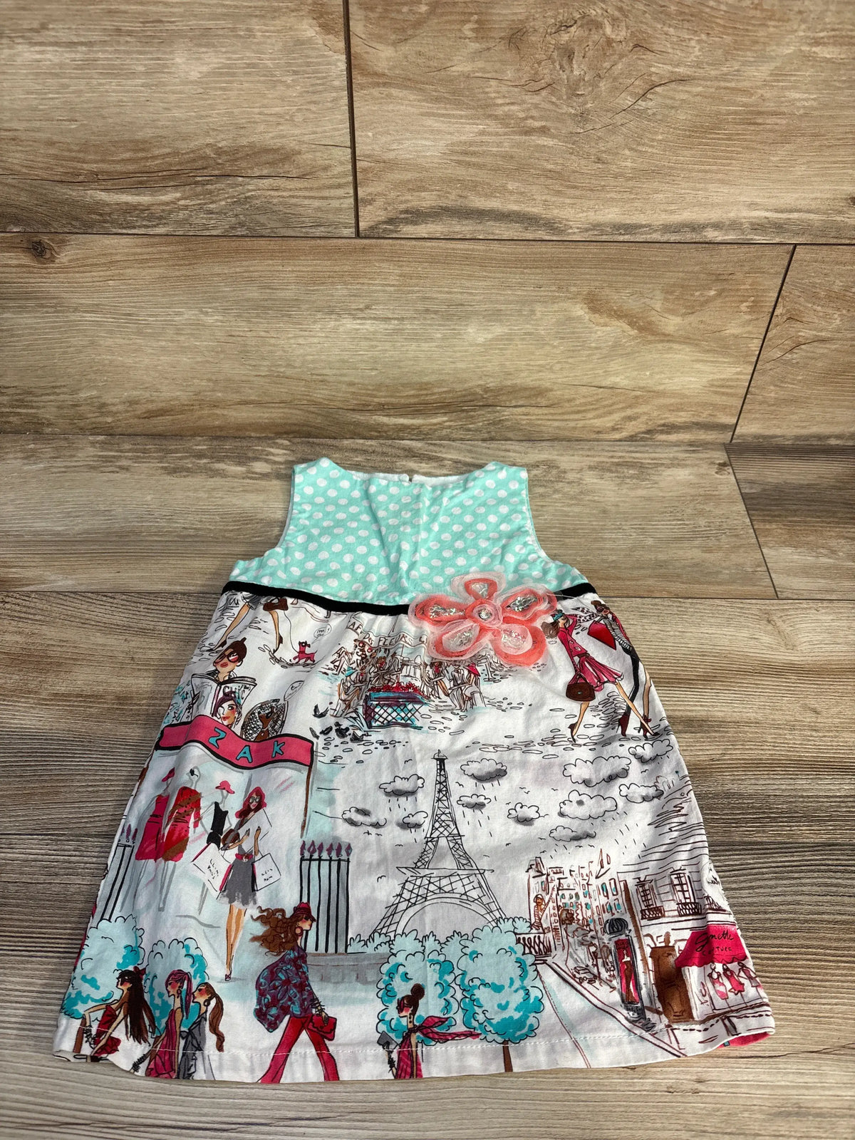 Front view of blue sleeveless dress showcasing a turquoise polka-dot bodice, Paris print skirt, and pink flower on wood floor
