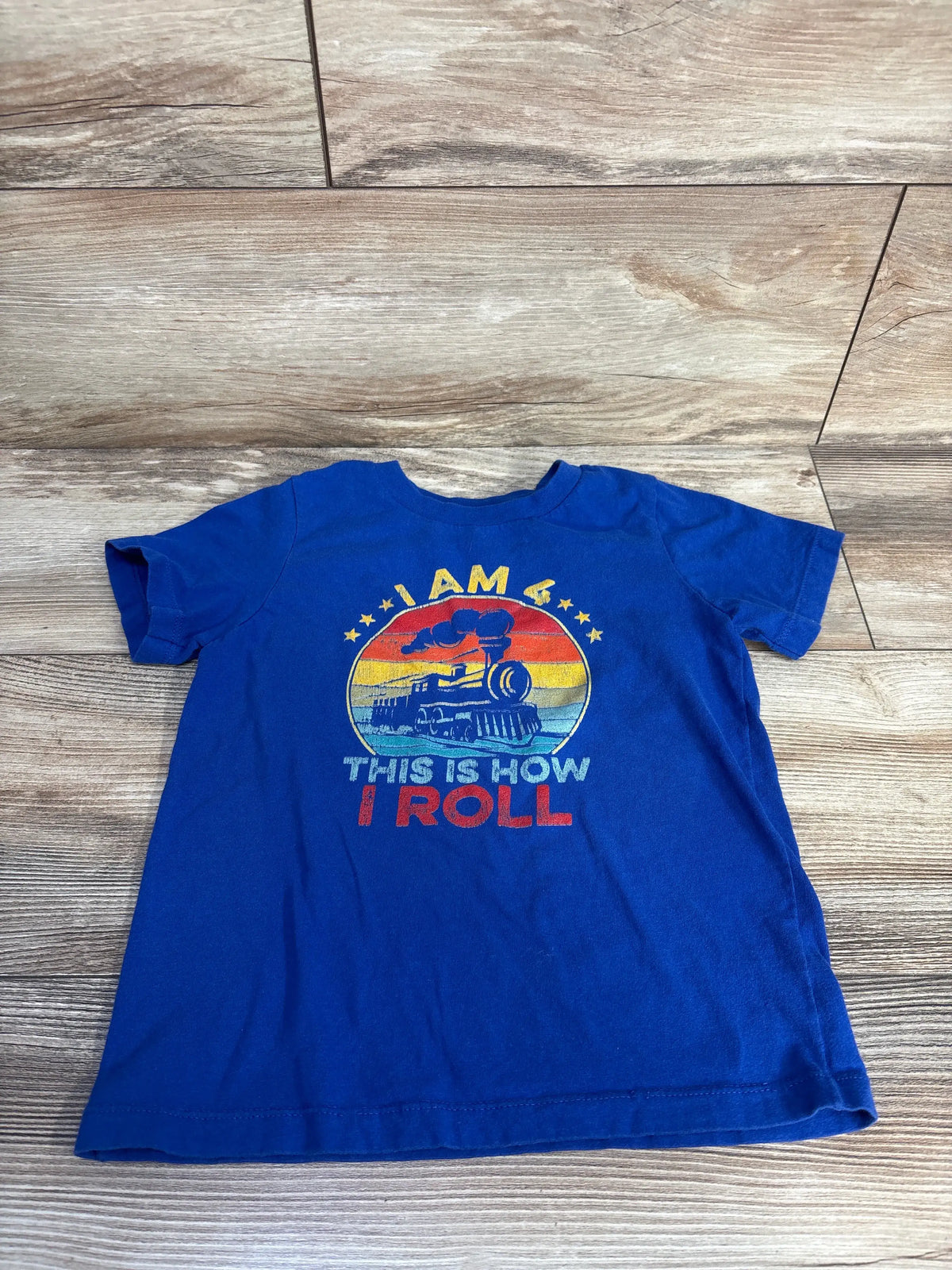 Front view of a blue Port & Company 4T toddler shirt with I AM 4 THIS IS HOW I ROLL train graphic on wood floor