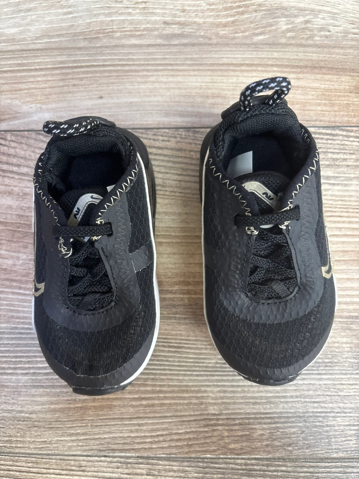 Top view of toddler Nike Air Max 2090 TD black sneakers showing metallic gold star accents and patterned heel pull tabs on wood floor