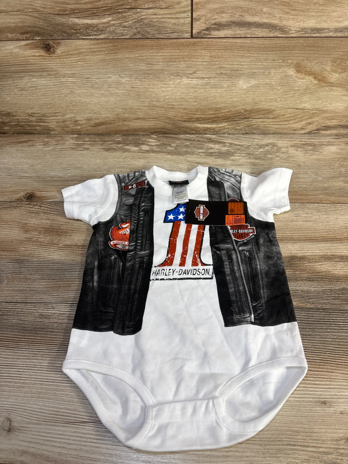 Flat lay of white Harley-Davidson baby bodysuit showing printed faux leather vest, American flag and Harley-Davidson logos on wood floor background