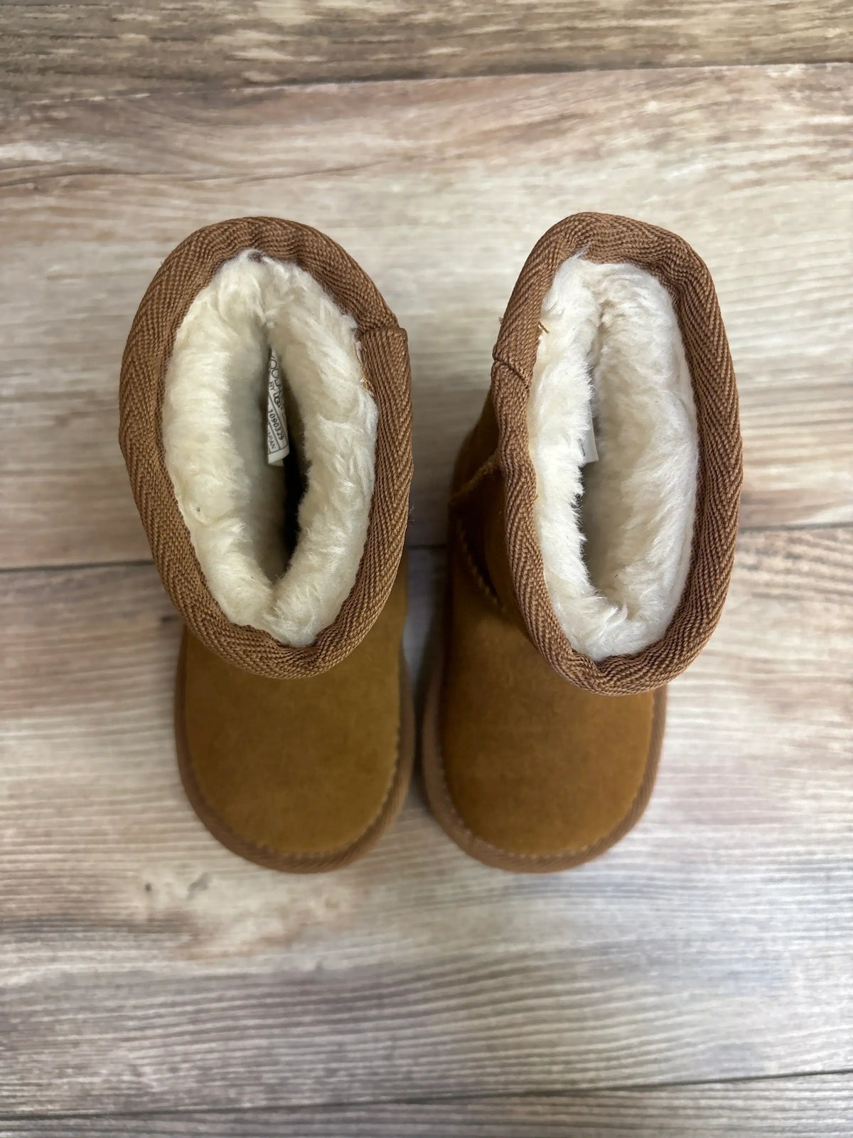 Top view of Koolaburra faux fur short boots showcasing chestnut brown suede uppers and cream fleece lined cuffs on wood plank floor