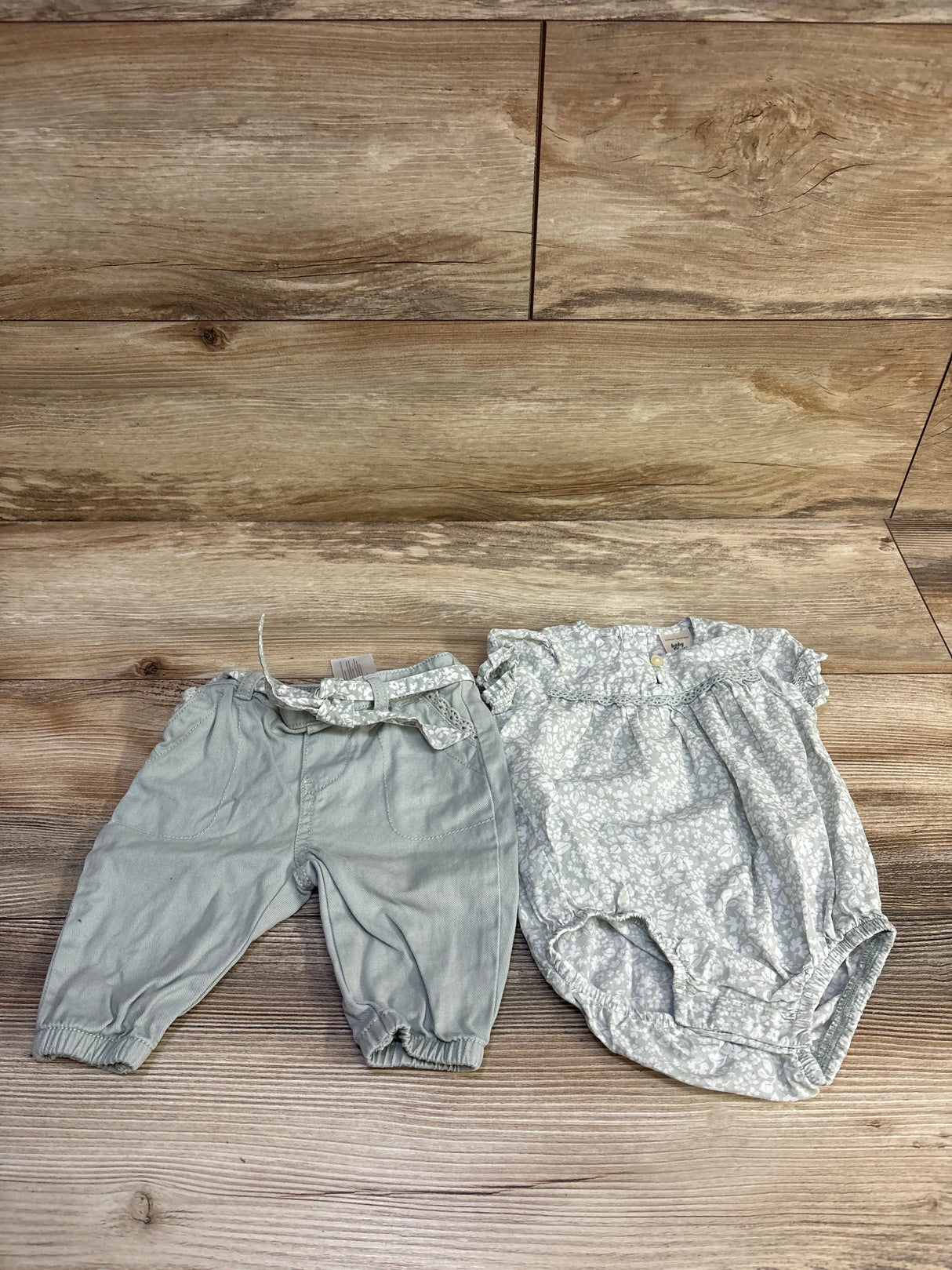Top-down view of Oshkosh 3-piece baby set size 3M: green pants, floral bubble romper, and fabric belt on wood floor