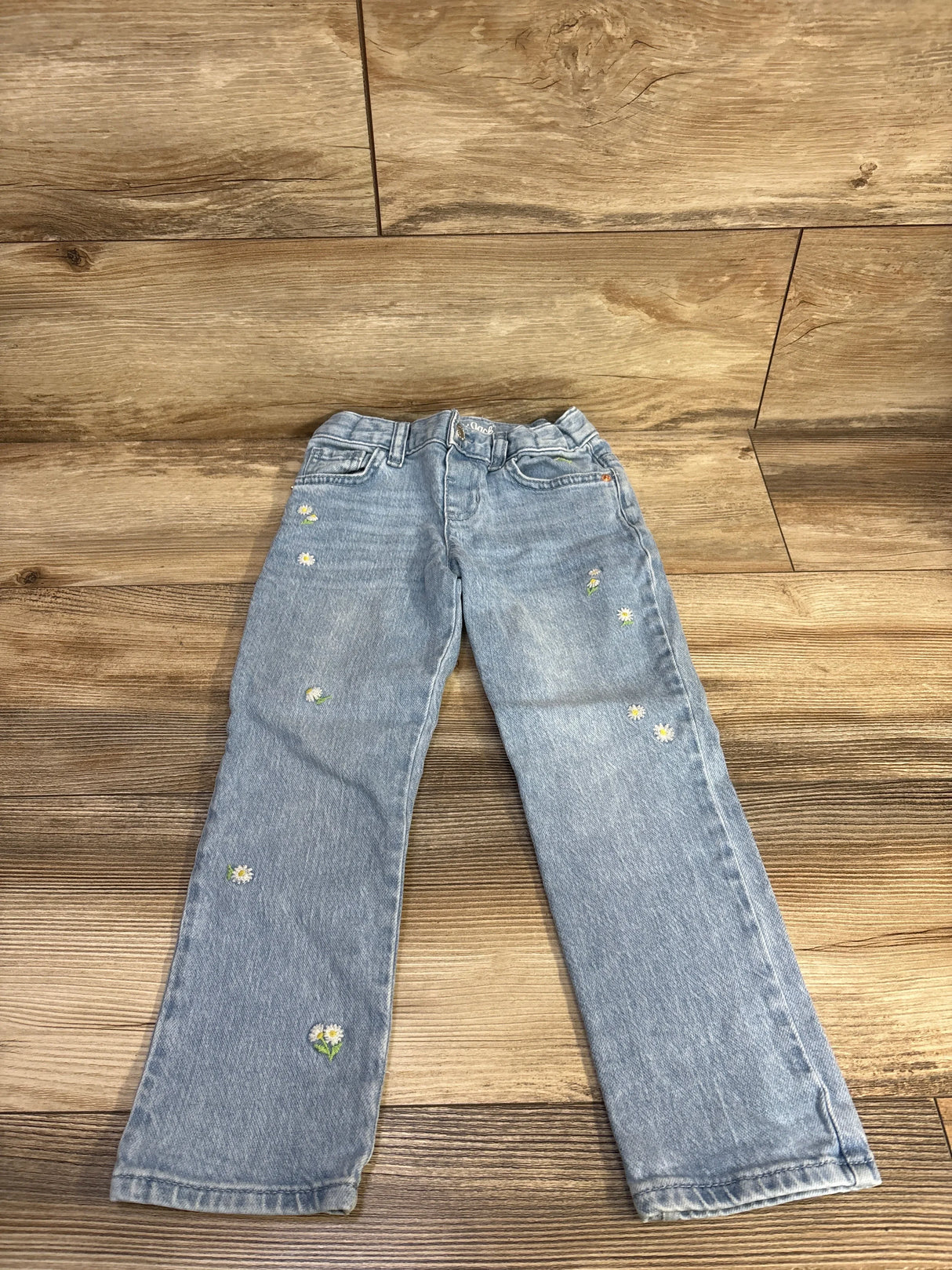 Front view of light blue toddler jeans size 5T showing embroidered white daisy motifs scattered on the legs on a wood floor background