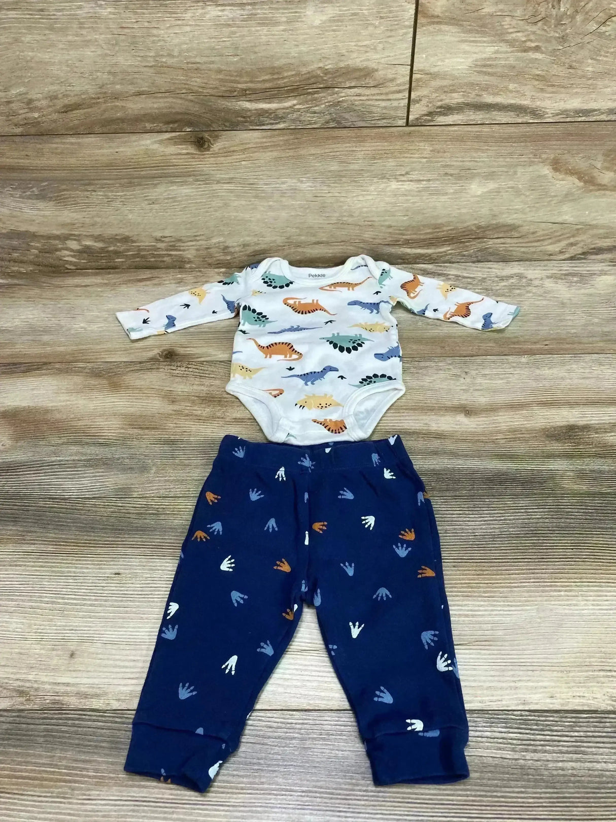 Top-down view of Pekkle 2-piece set: white long-sleeve dinosaur bodysuit and navy footprint pants on wood floor