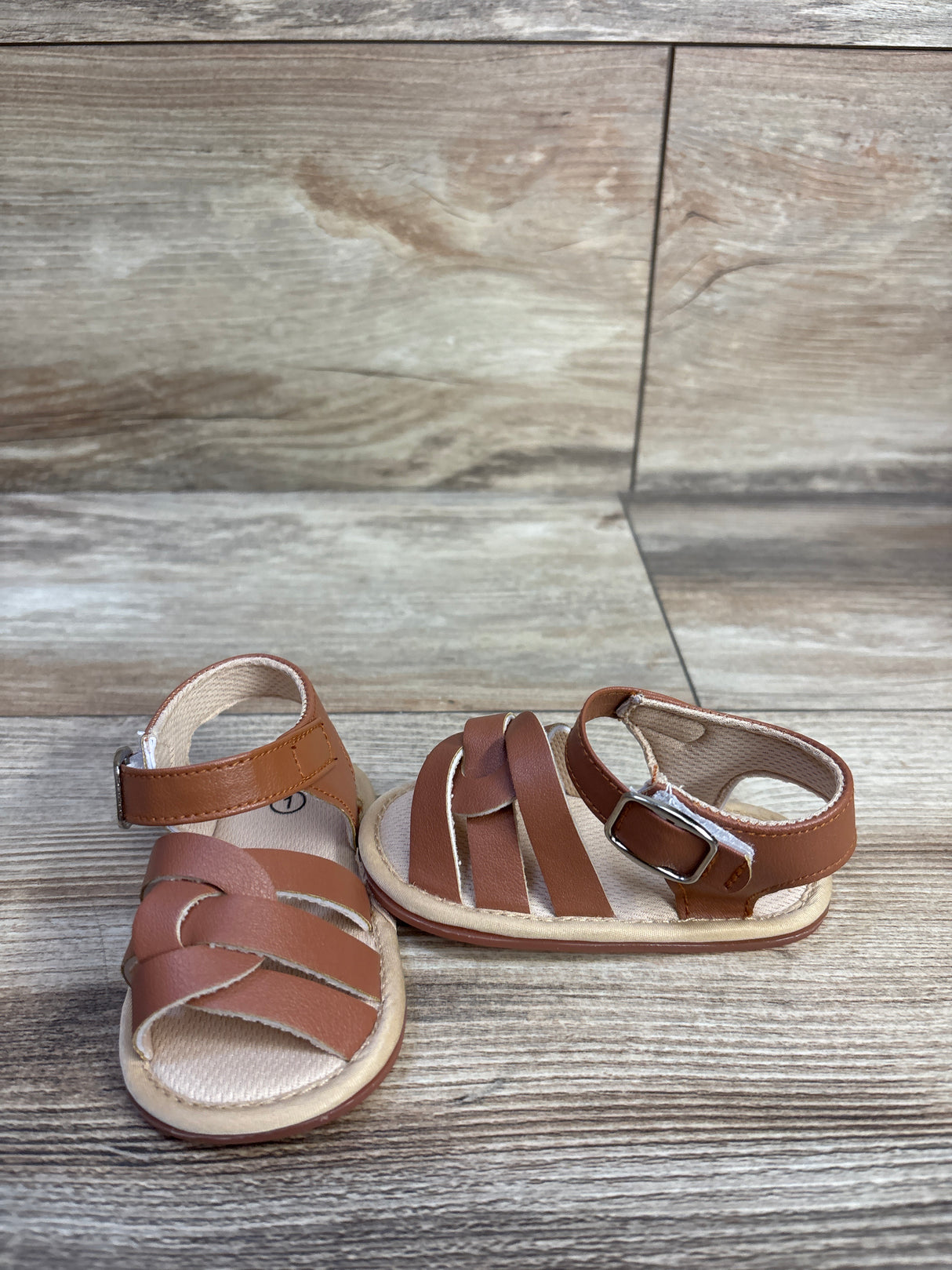 Three-quarter view of brown Hsdsbebe soft sole baby sandals showing braided front straps and ankle strap with buckle on wood background