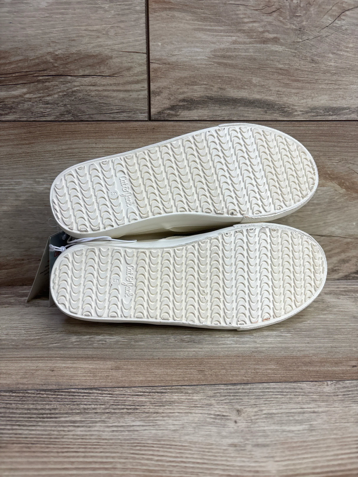 Bottom view of white rubber-soled toddler shoes with textured grip, size 5, on wood floor